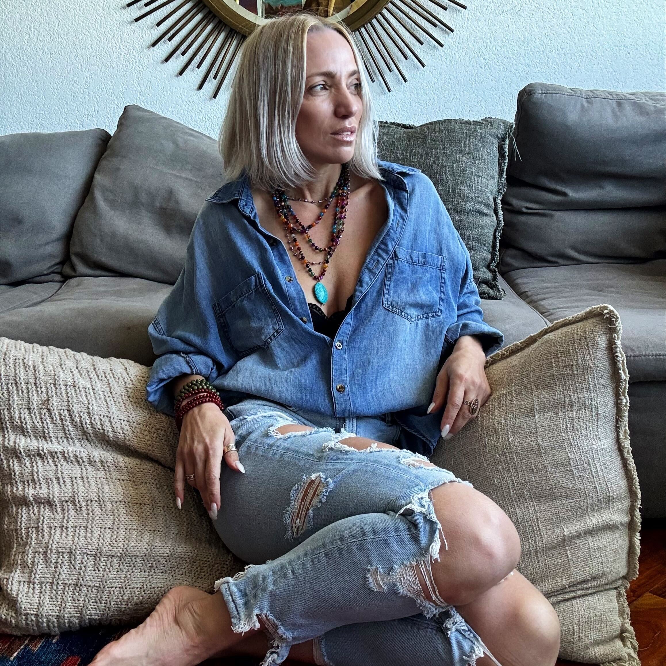 Woman sitting on a couch wearing a blue denim shirt and ripped jeans, with a decorative wall mirror in the background.