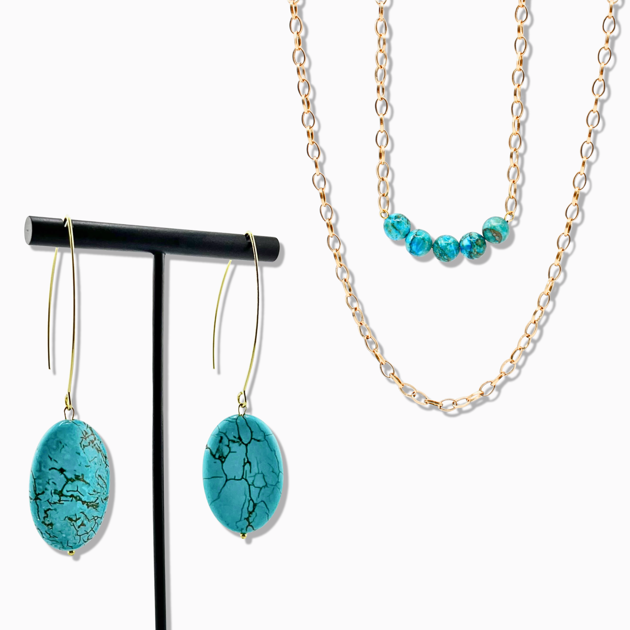 Turquoise earrings on a black stand with a gold chain necklace on a white background