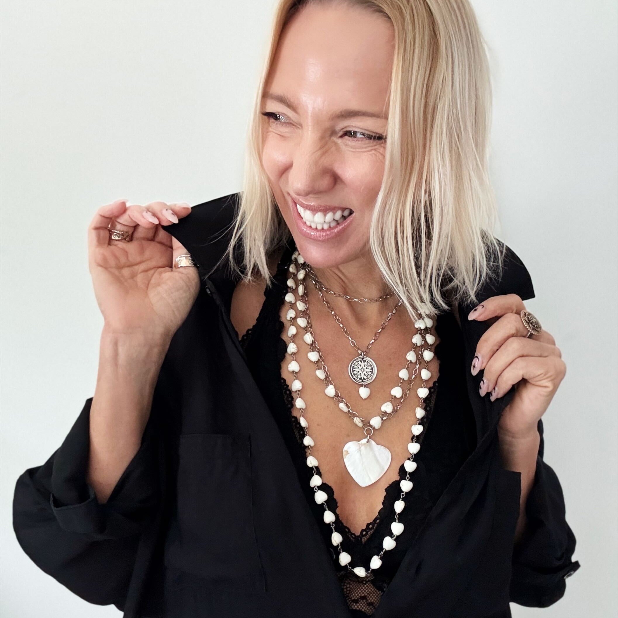 A smiling person with blonde hair wears a black shirt and layered necklaces, including a delicate layering chain adorned with howlite heart beads, against a plain background.