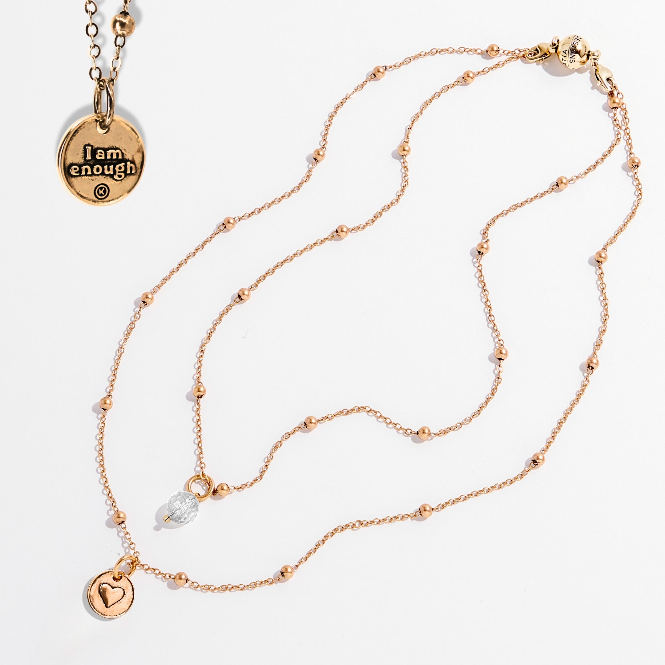 Two dainty necklaces with pendants, one with a silver finish and the other with a gold finish. Both have a chain link design and feature a circular pendant with the phrase 'I am enough' and a heart symbol respectively.