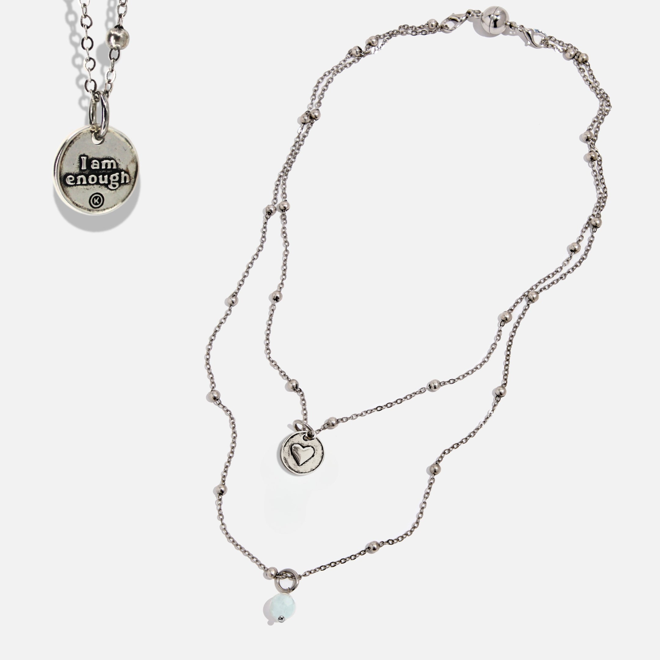 Two dainty necklaces with pendants, one with a silver finish and the other with a gold finish. Both have a chain link design and feature a circular pendant with the phrase 'I am enough' and a heart symbol respectively.