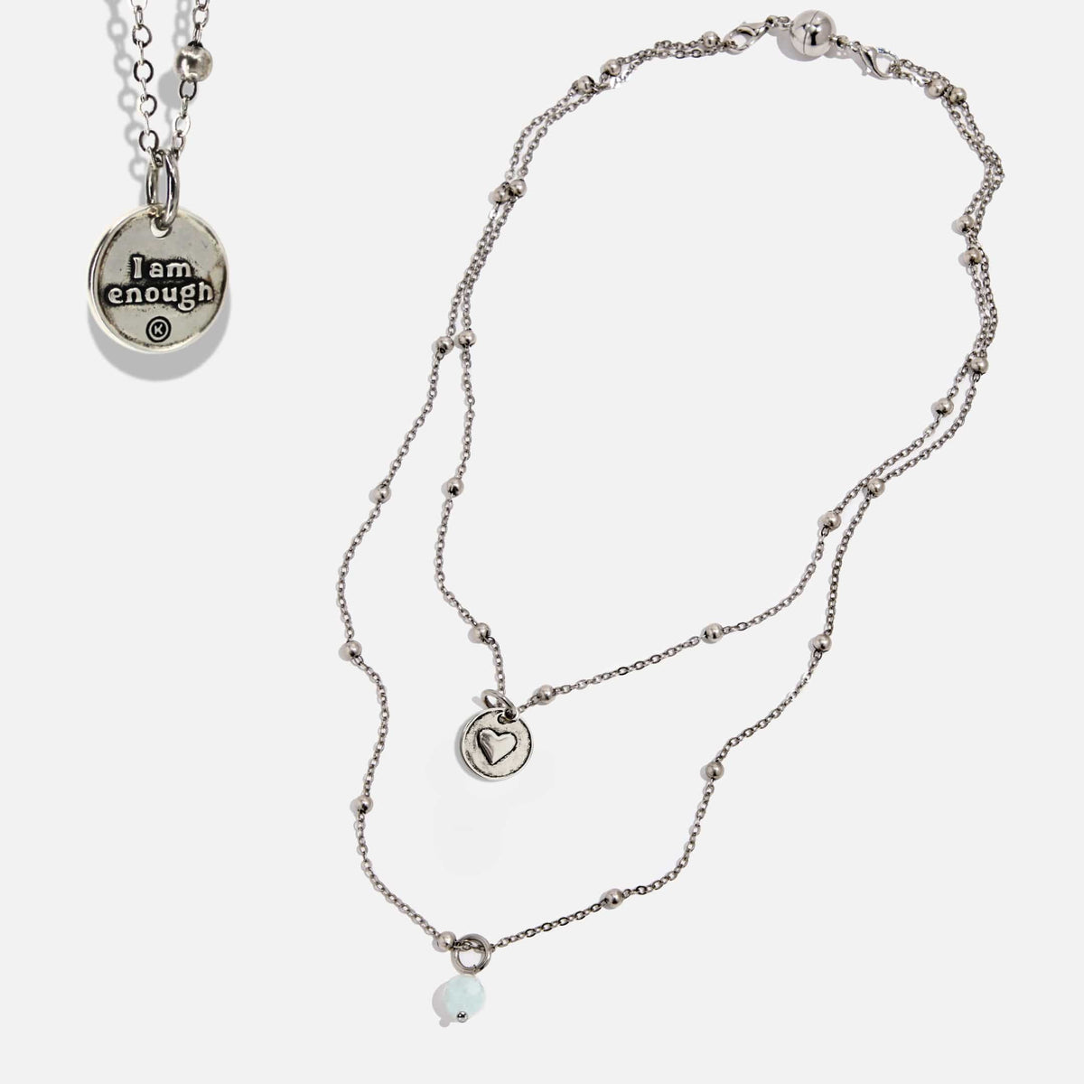 Two dainty necklaces with pendants, one with a silver finish and the other with a gold finish. Both have a chain link design and feature a circular pendant with the phrase 'I am enough' and a heart symbol respectively.