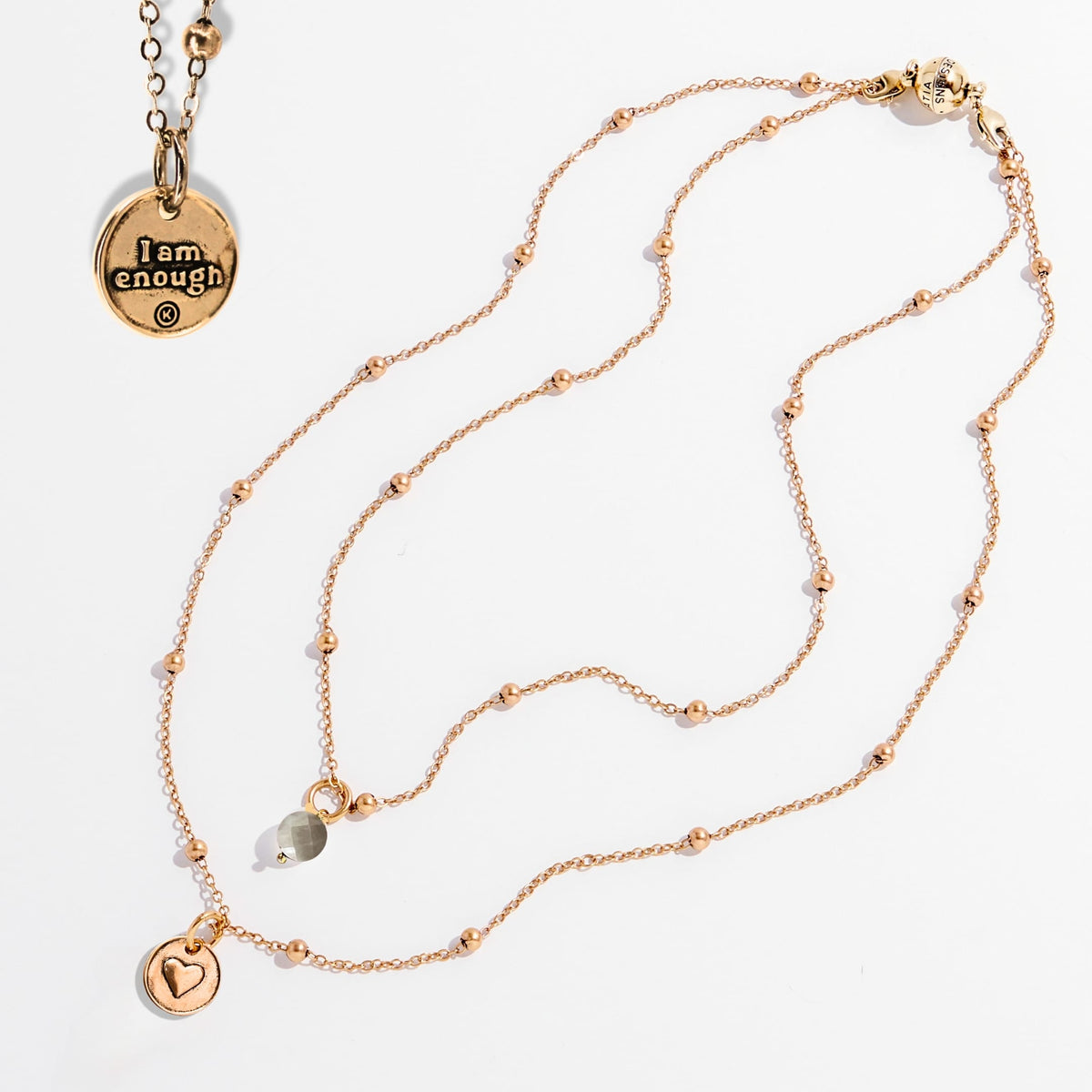 Two dainty necklaces with pendants, one with a silver finish and the other with a gold finish. Both have a chain link design and feature a circular pendant with the phrase 'I am enough' and a heart symbol respectively.