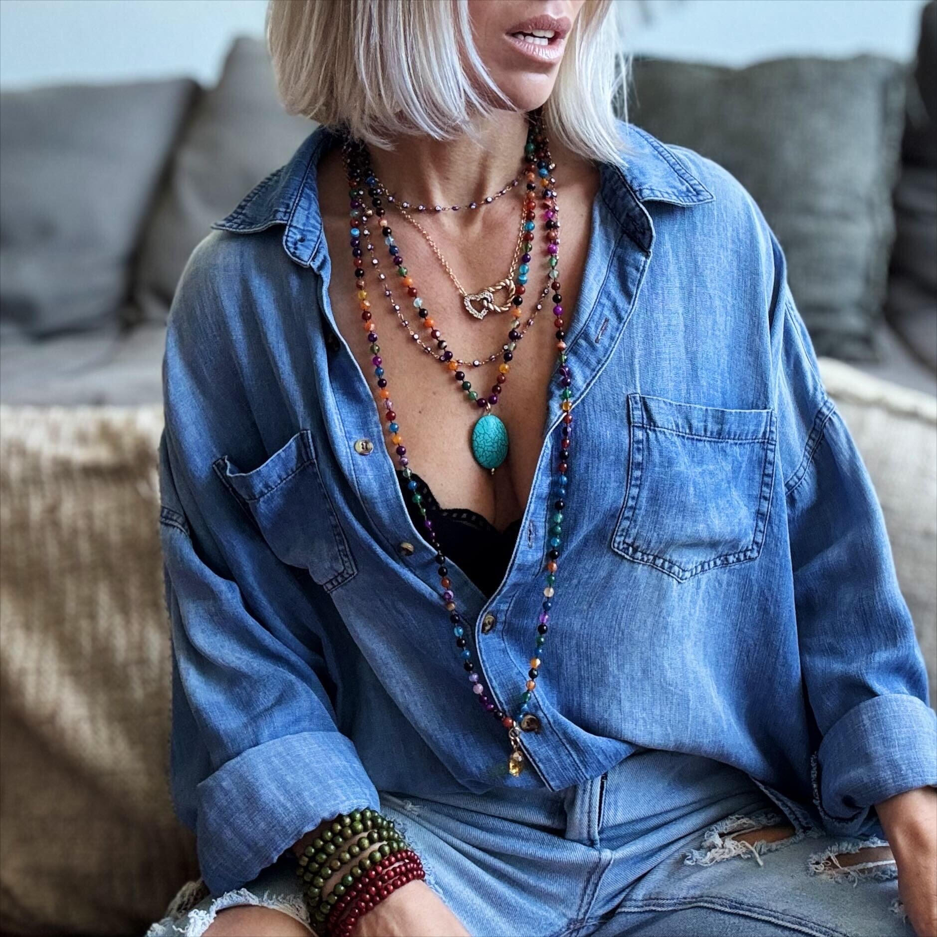 Person wearing a blue denim shirt with layered necklaces and bracelets in a casual setting.