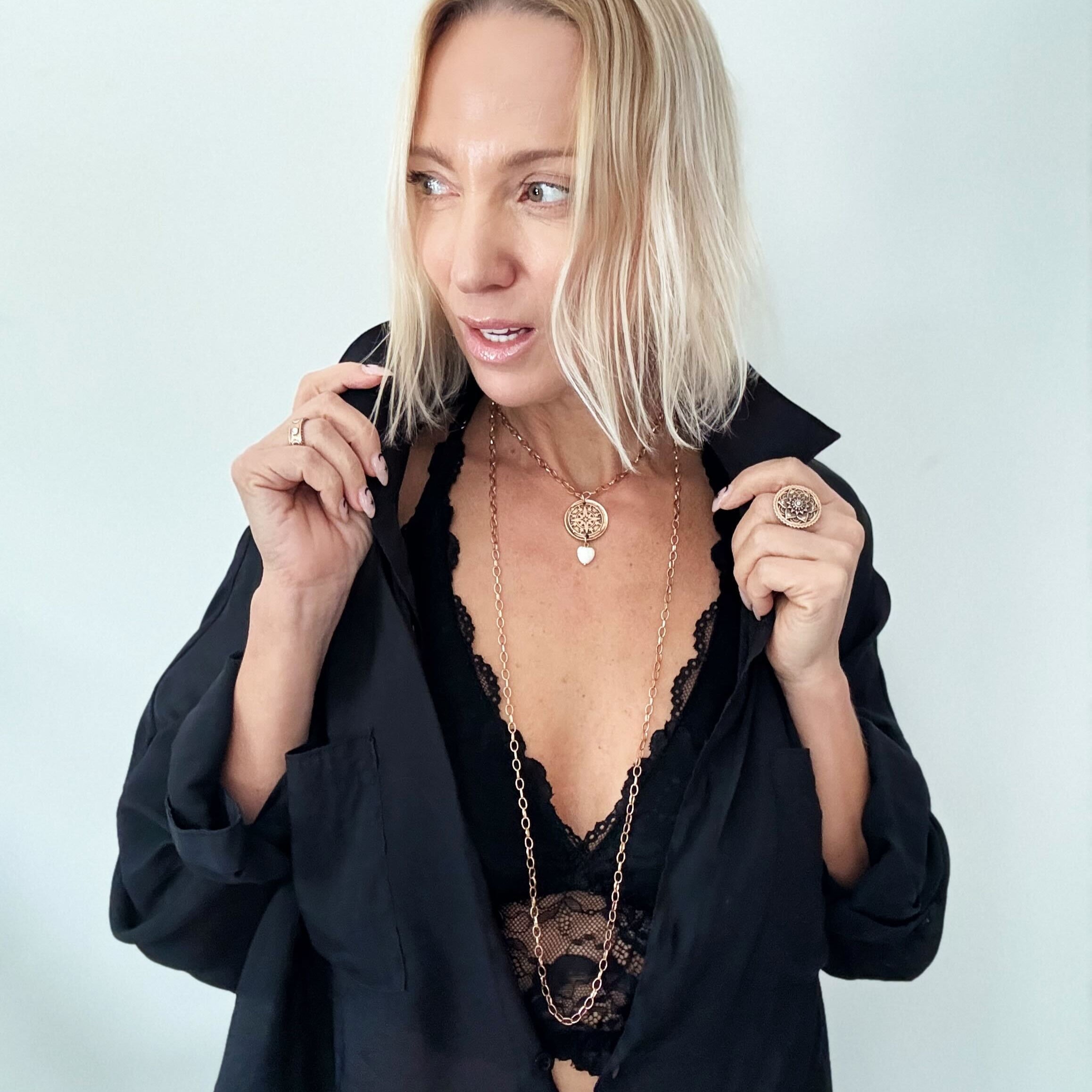 A smiling person with blonde hair wears a black shirt and layered necklaces, including a delicate layering chain adorned with howlite heart beads, against a plain background.