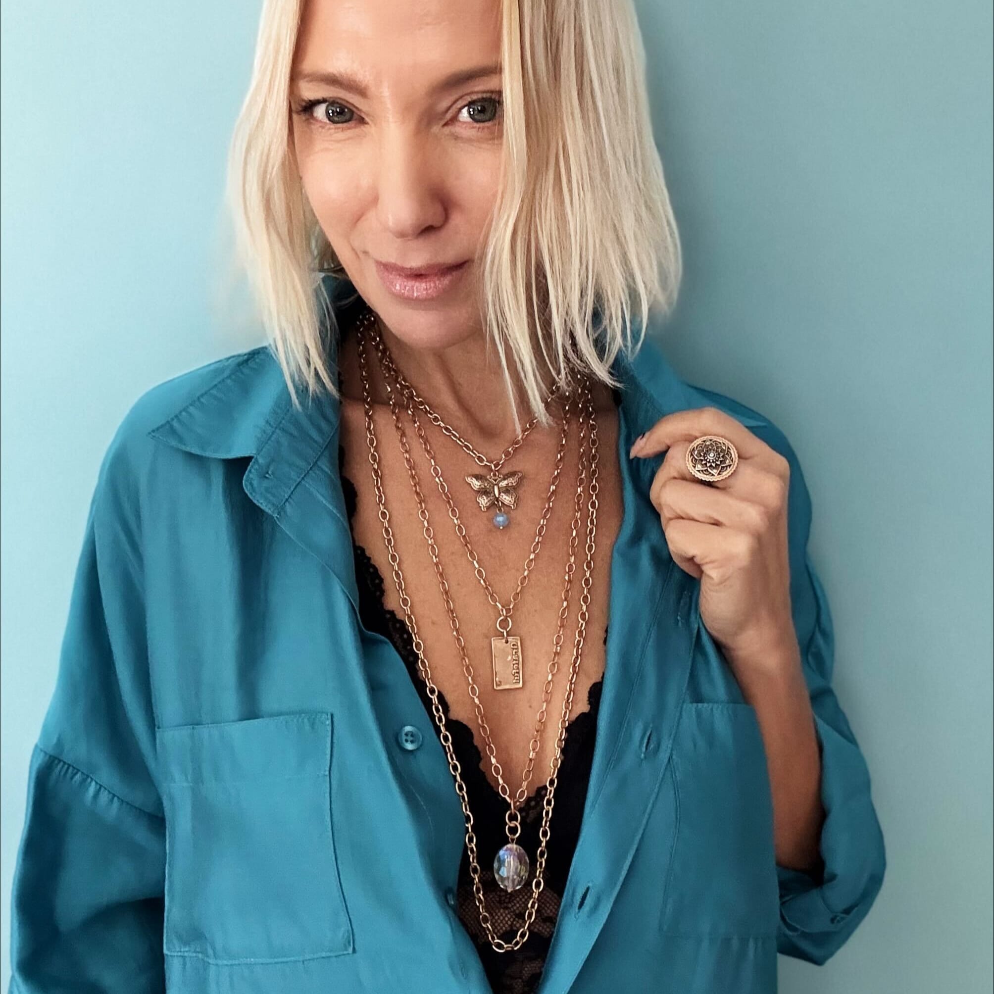 Woman in teal shirt with tousled blonde bob, wearing a butterfly pendant and large ring, against a blue background.