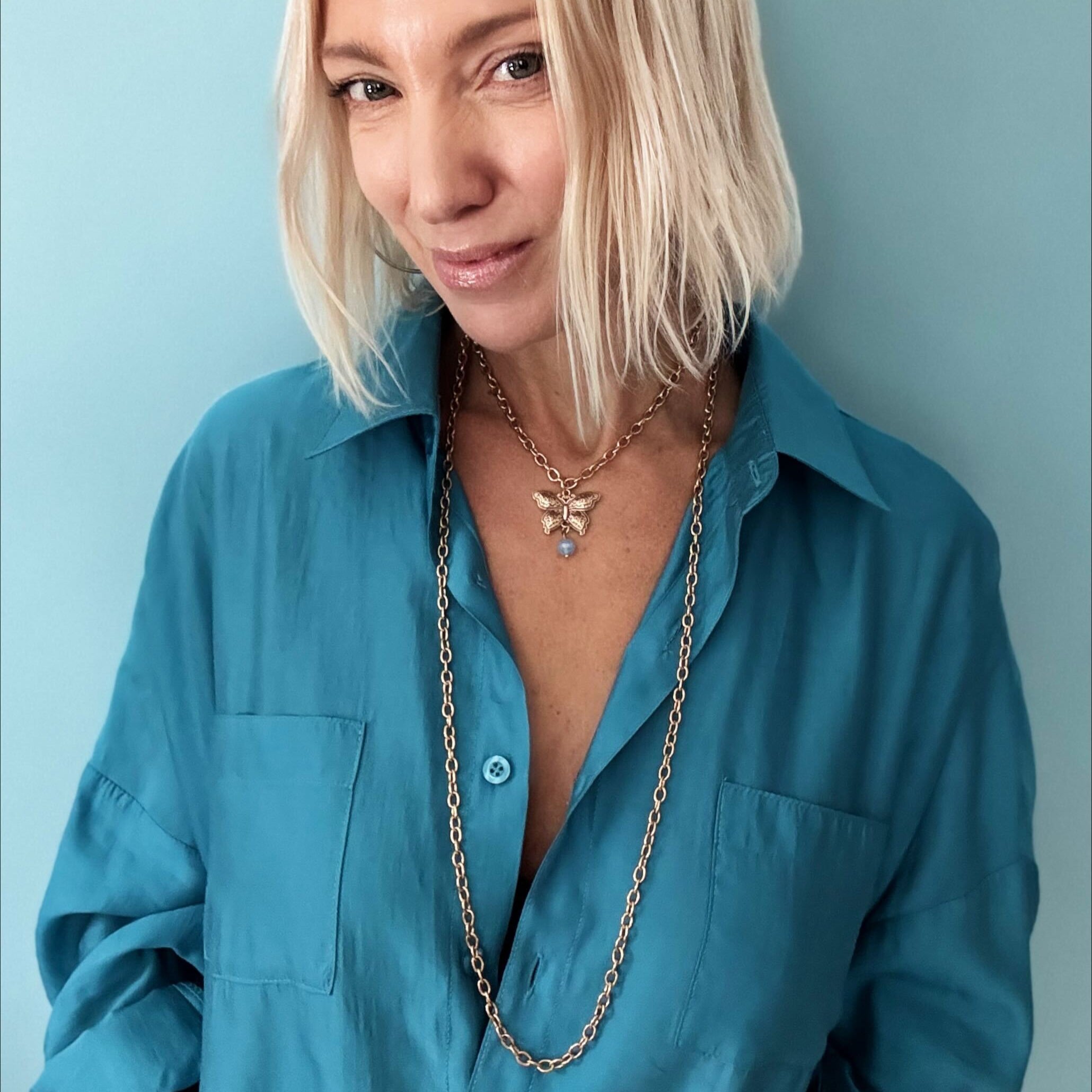 Woman in teal shirt with tousled blonde bob, wearing a butterfly pendant and large ring, against a blue background.