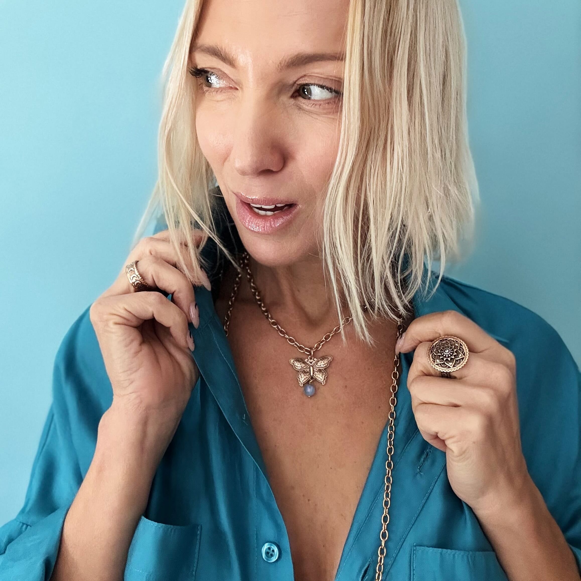 Woman in teal shirt with tousled blonde bob, wearing a butterfly pendant and large ring, against a blue background.