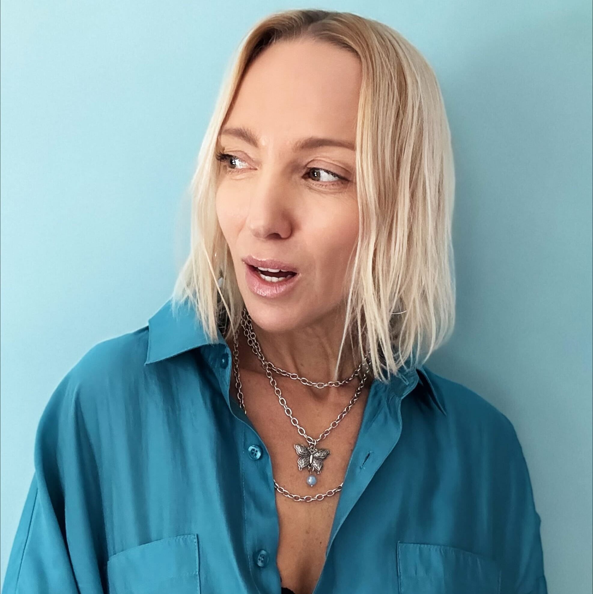 Woman in teal shirt with tousled blonde bob, wearing a butterfly pendant and large ring, against a blue background.