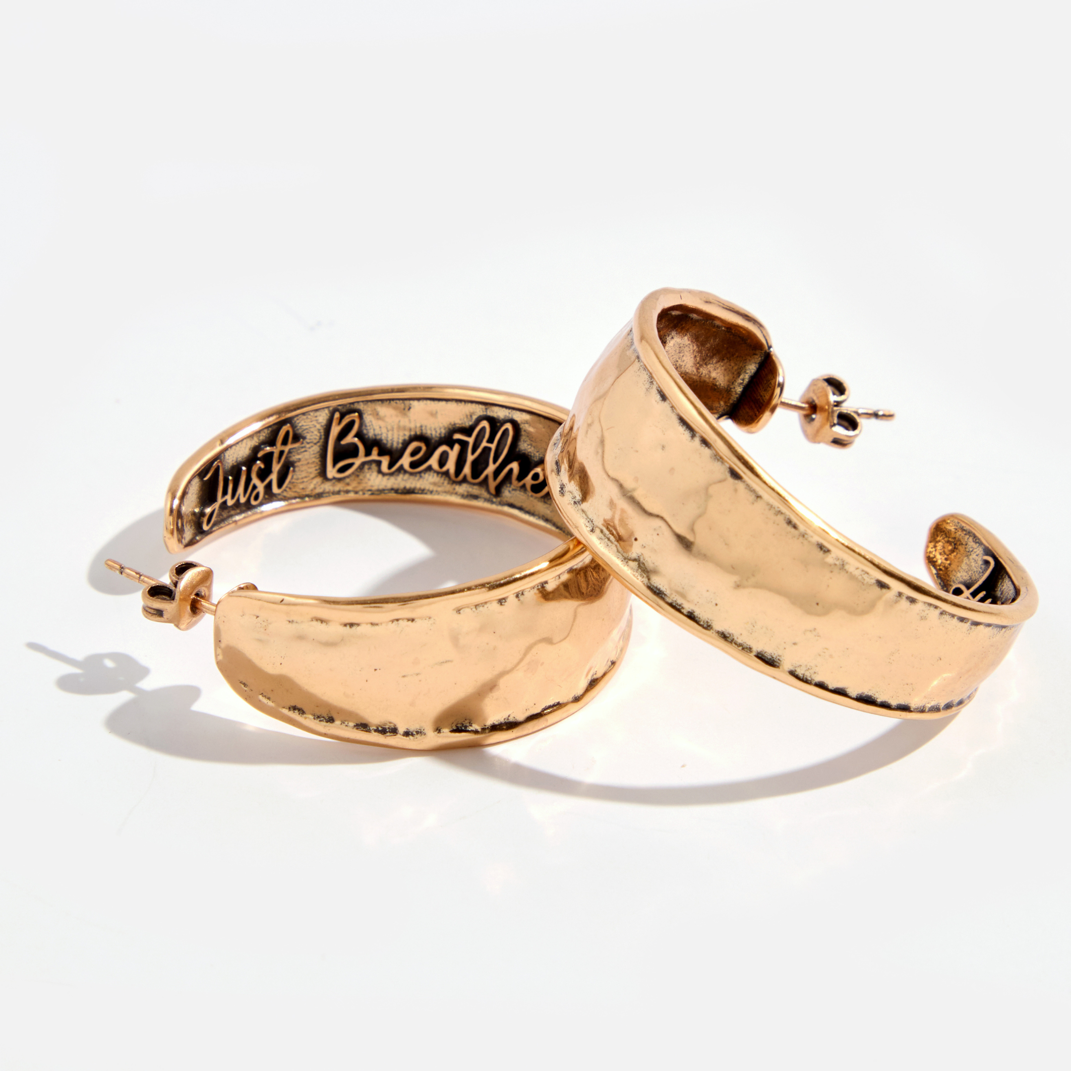 A pair of hoop earrings with a hammered texture, one with 'Just Breathe' inscribed, in silver and gold colors.