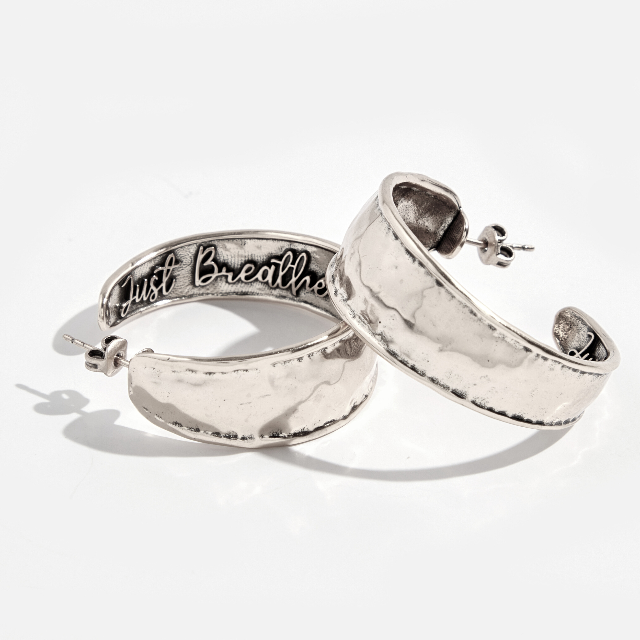 A pair of hoop earrings with a hammered texture, one with 'Just Breathe' inscribed, in silver and gold colors.