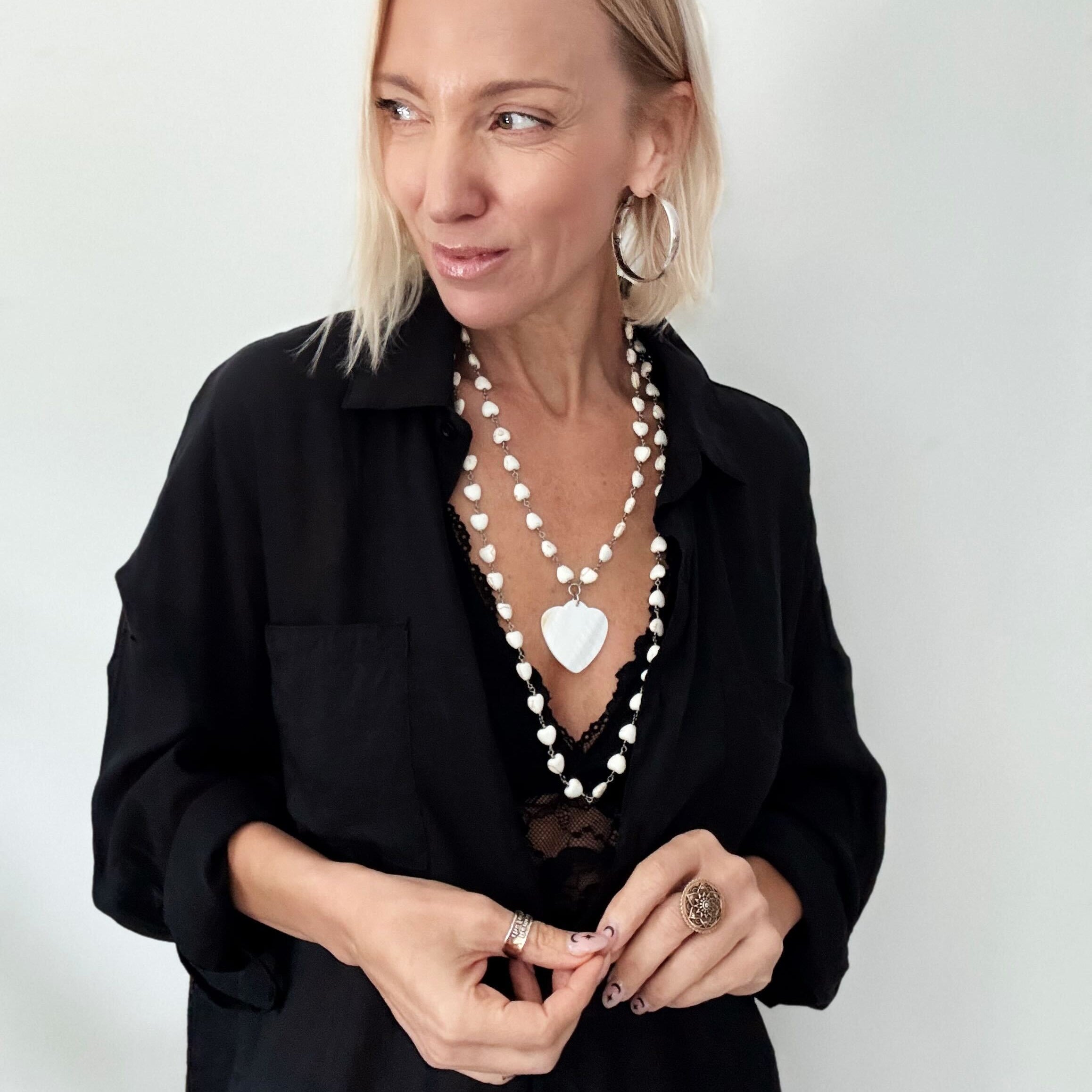 A smiling person with blonde hair wears a black shirt and layered necklaces, including a delicate layering chain adorned with howlite heart beads, against a plain background.