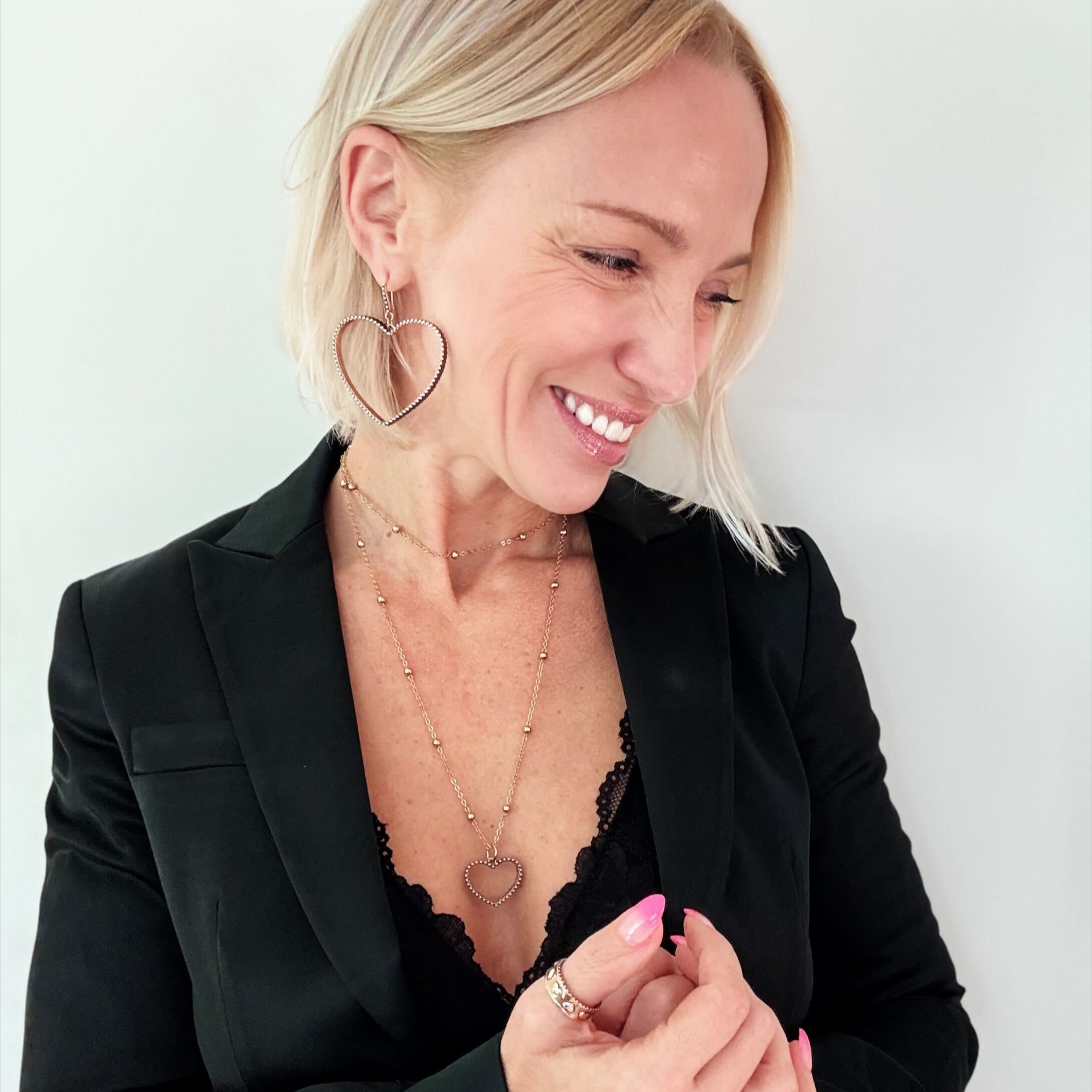 A person with short blonde hair is smiling and looking down. They are wearing a black jacket over a lace top, large open heart pendant earrings, and layered mixed-metal necklaces.