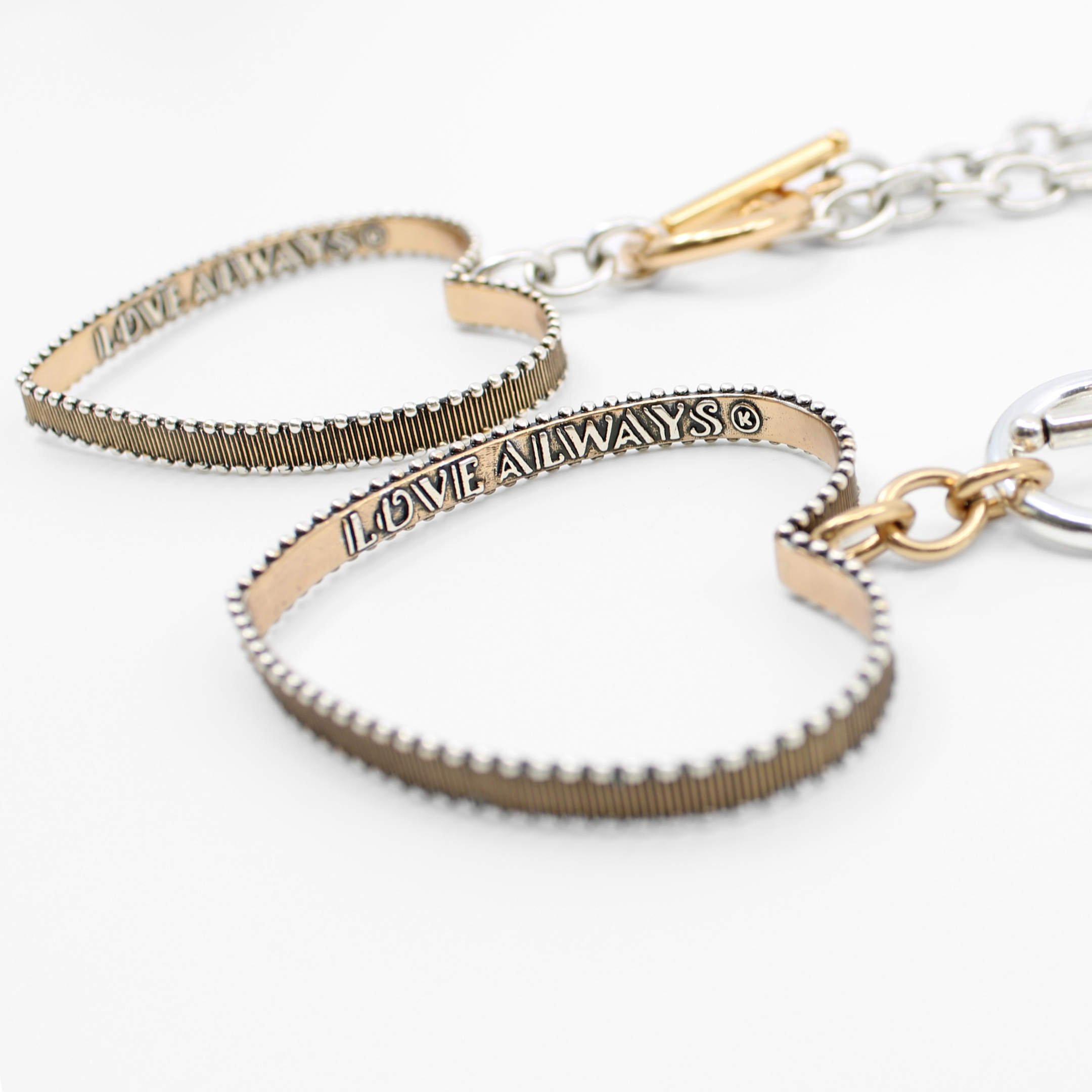 Close-up of two heart-shaped PENDANTS with textured edges, connected by a toggle closure. Each heart bears the inscription LOVE ALWAYS. They are placed on a white background.