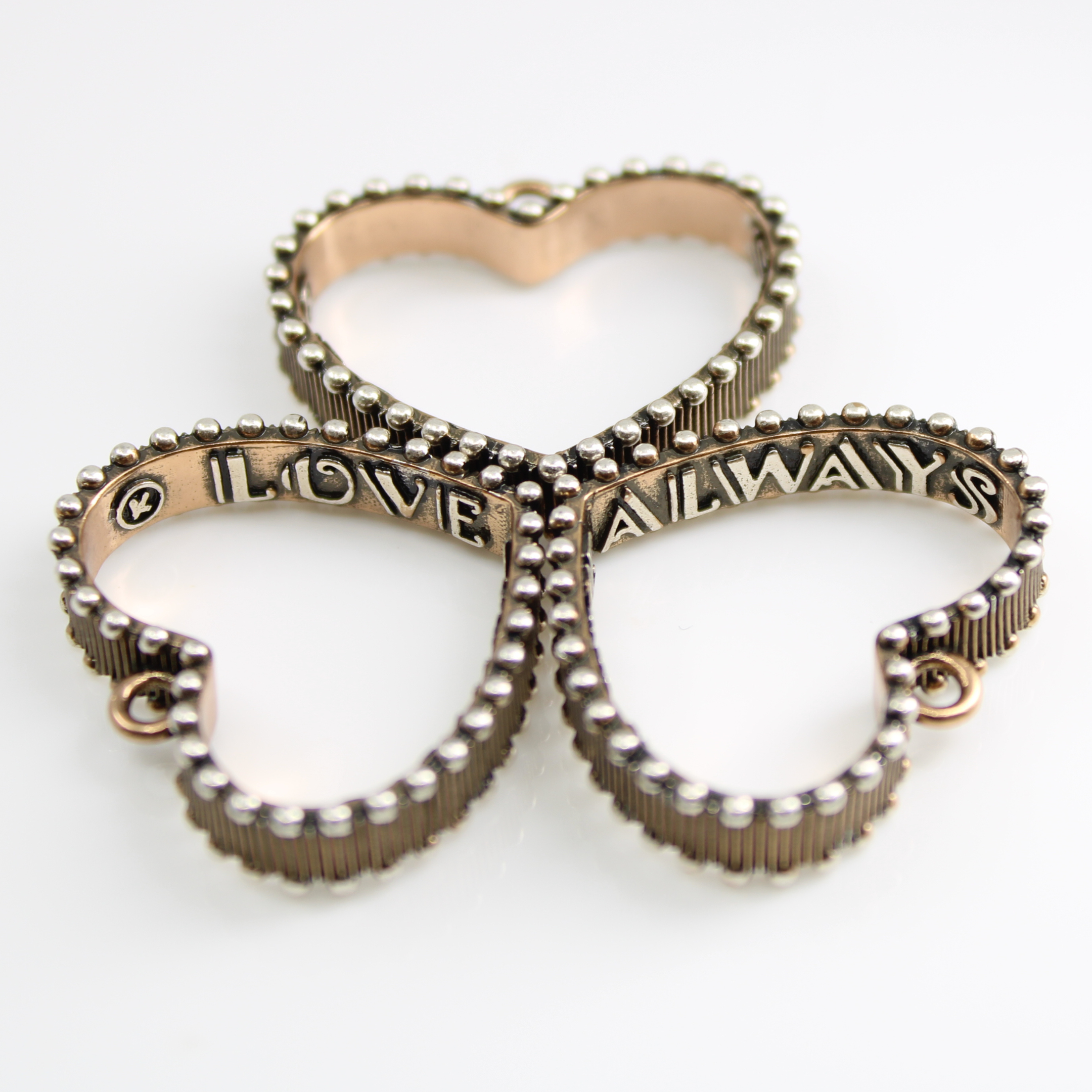 A mixed-metal pendant DISPLAYING three hearts, each outlined with a scalloped trim design. LOVE ALWAYS is inscribed inside the hearts. The open heart pendant hangs gracefully, accentuated by a secure magnetic clasp on a white background.