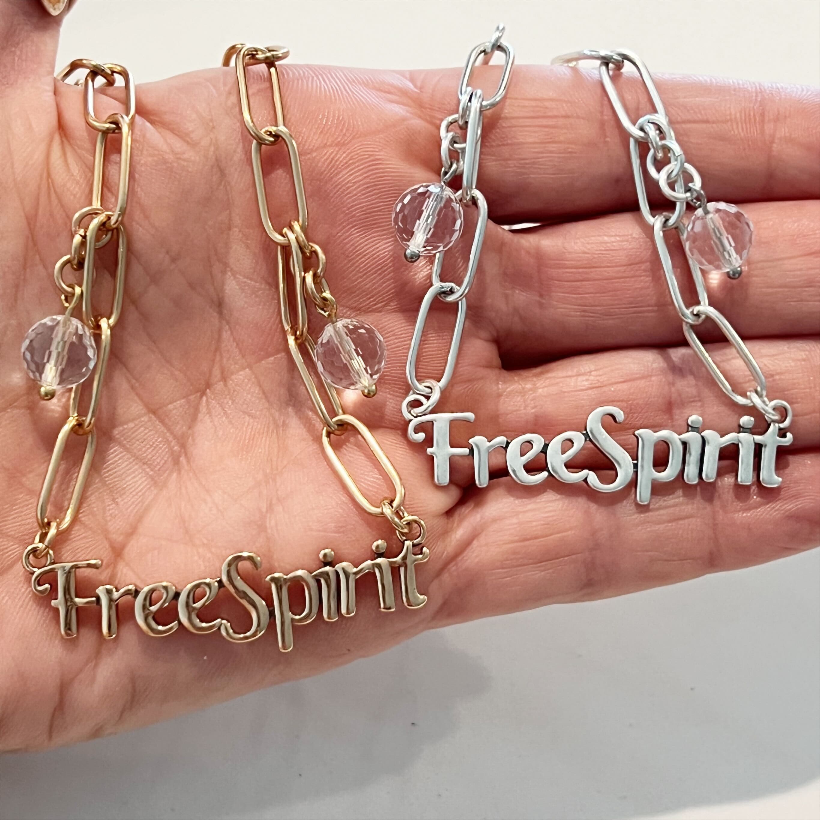 Two long chain necklaces, one plated in gold and the other in silver, each with the text 'FreeSpirit' and crystal quartz accents laid out on a hand.