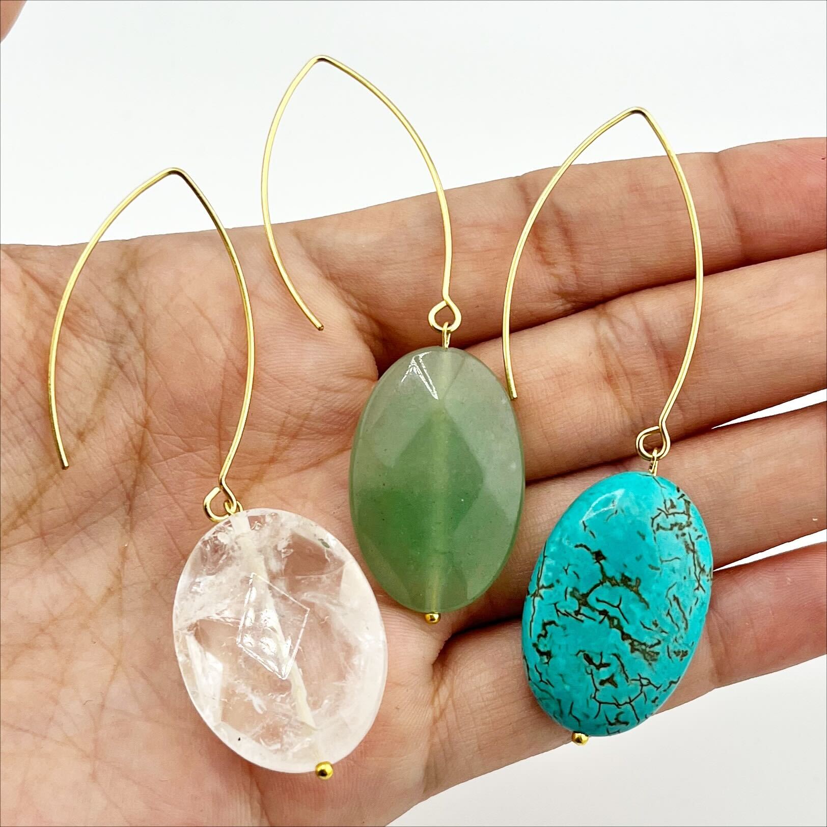 Three stone drop earrings in gold finish, each with a different color of stone: clear quartz, green aventurine, and blue howlite, held in a person's hand.