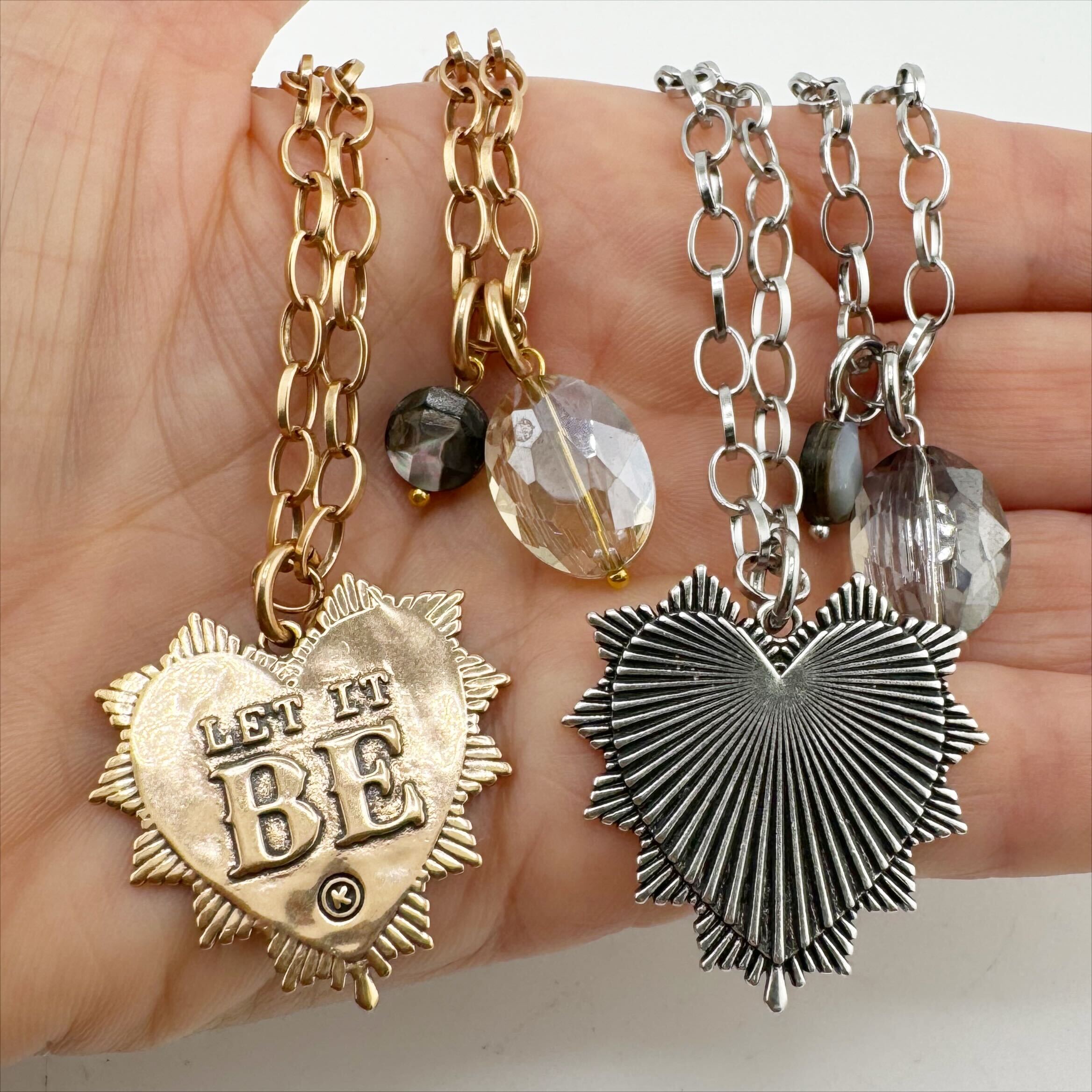 Two necklaces, one with a gold-plated heart pendant and the other with a silver-plated heart pendant, each featuring a faceted crystal accent and a black lip pearl on a chain, held in a person's hand.