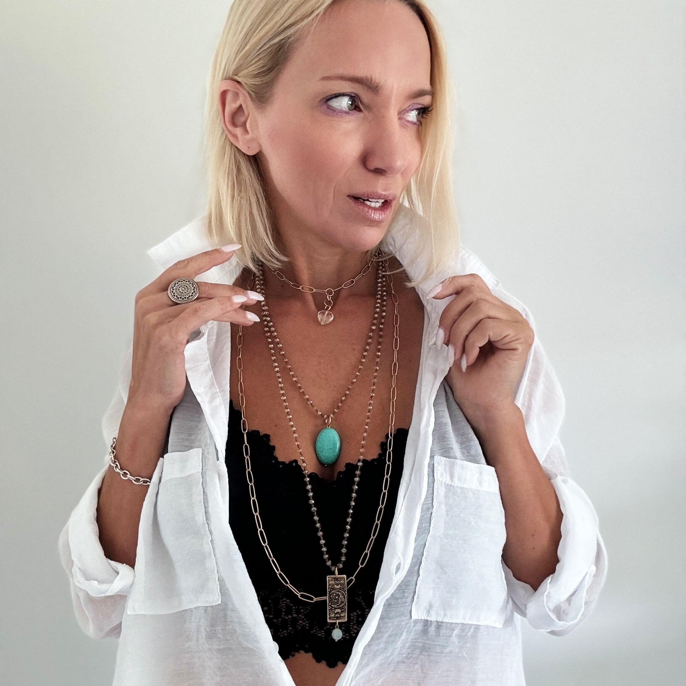 A person wearing a white shirt and black lace top, showcasing layered necklaces with silver and gold tones, crystal beads, and pendant charms.