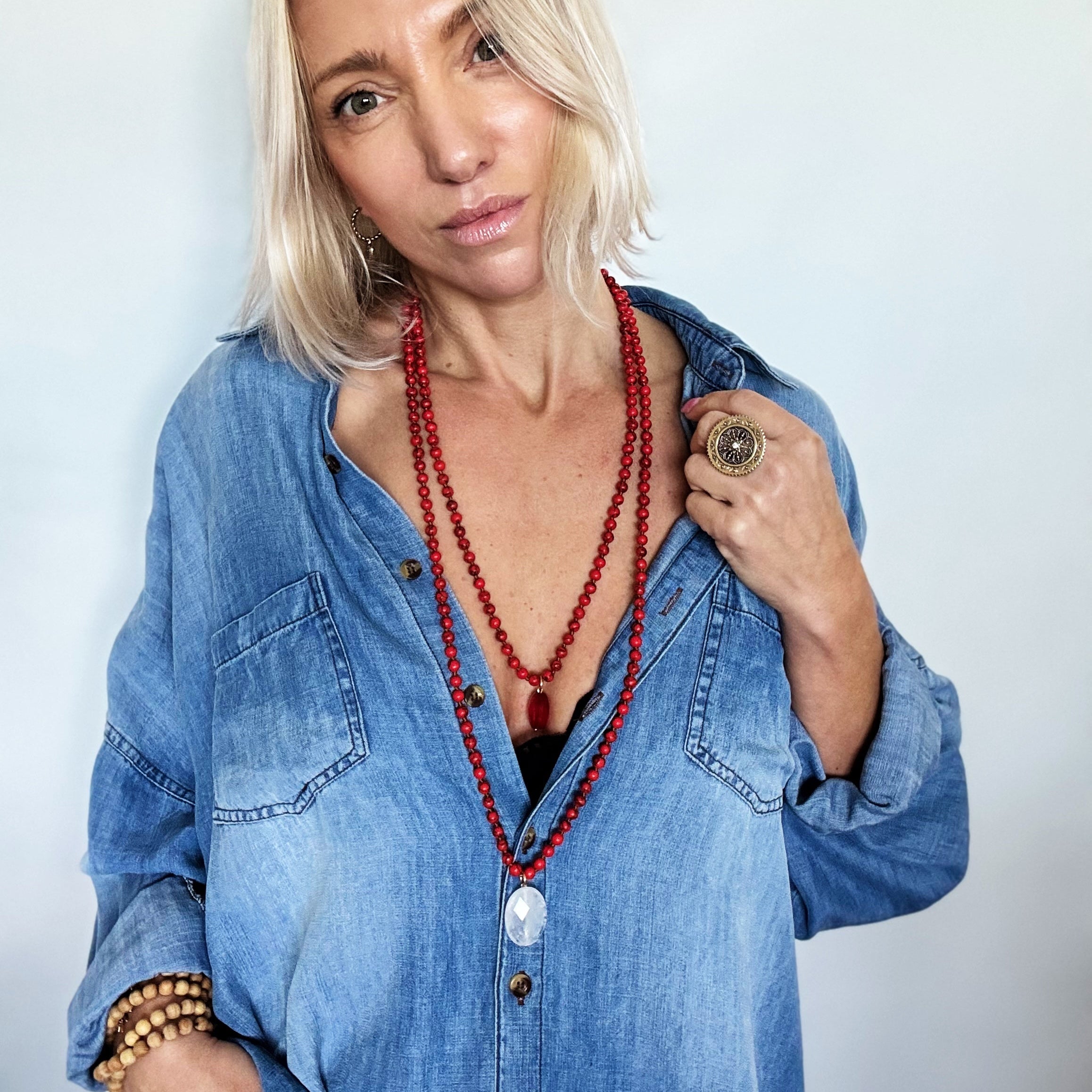 Woman wearing a blue denim shirt and red beaded necklace against a plain background