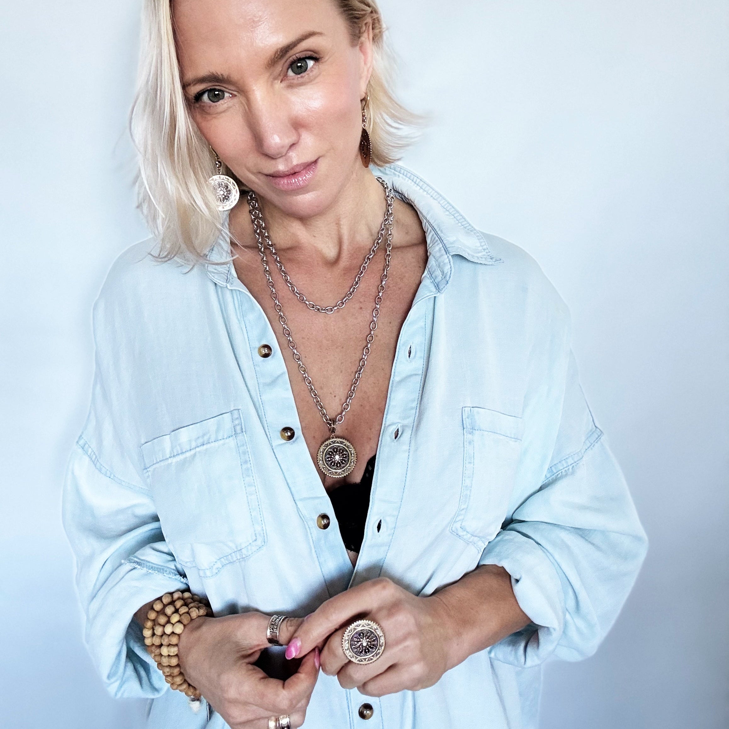 Woman wearing a light blue denim shirt with layered necklaces and jewelry against a plain background