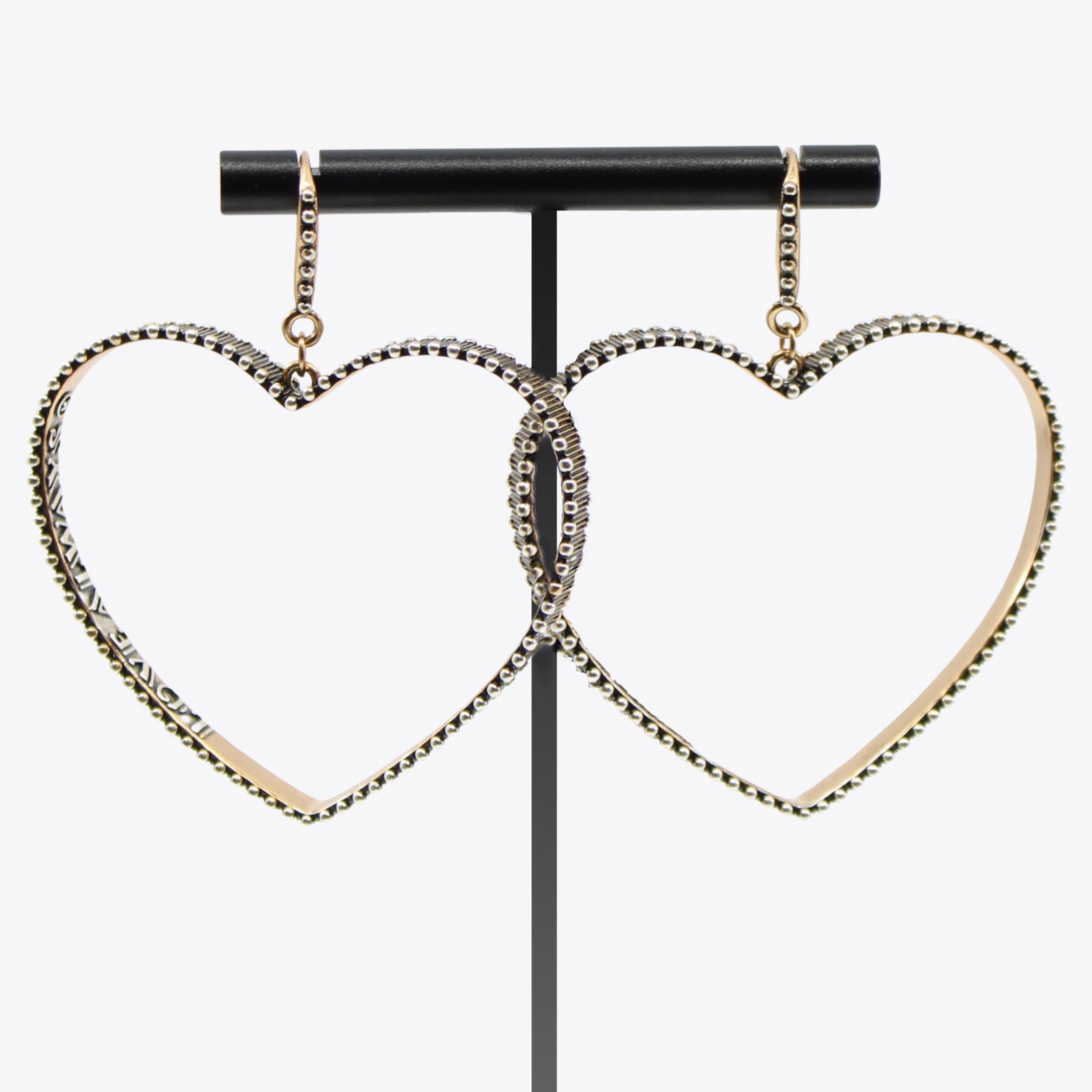 Two large, heart-shaped drop earrings from Katia Designs, with studded embellishments, make a bold statement as they gleam on a black stand against a plain white background. These open-heart pieces embody the essence of love and mix-metal elegance. Love Always message inscribed on the inside.