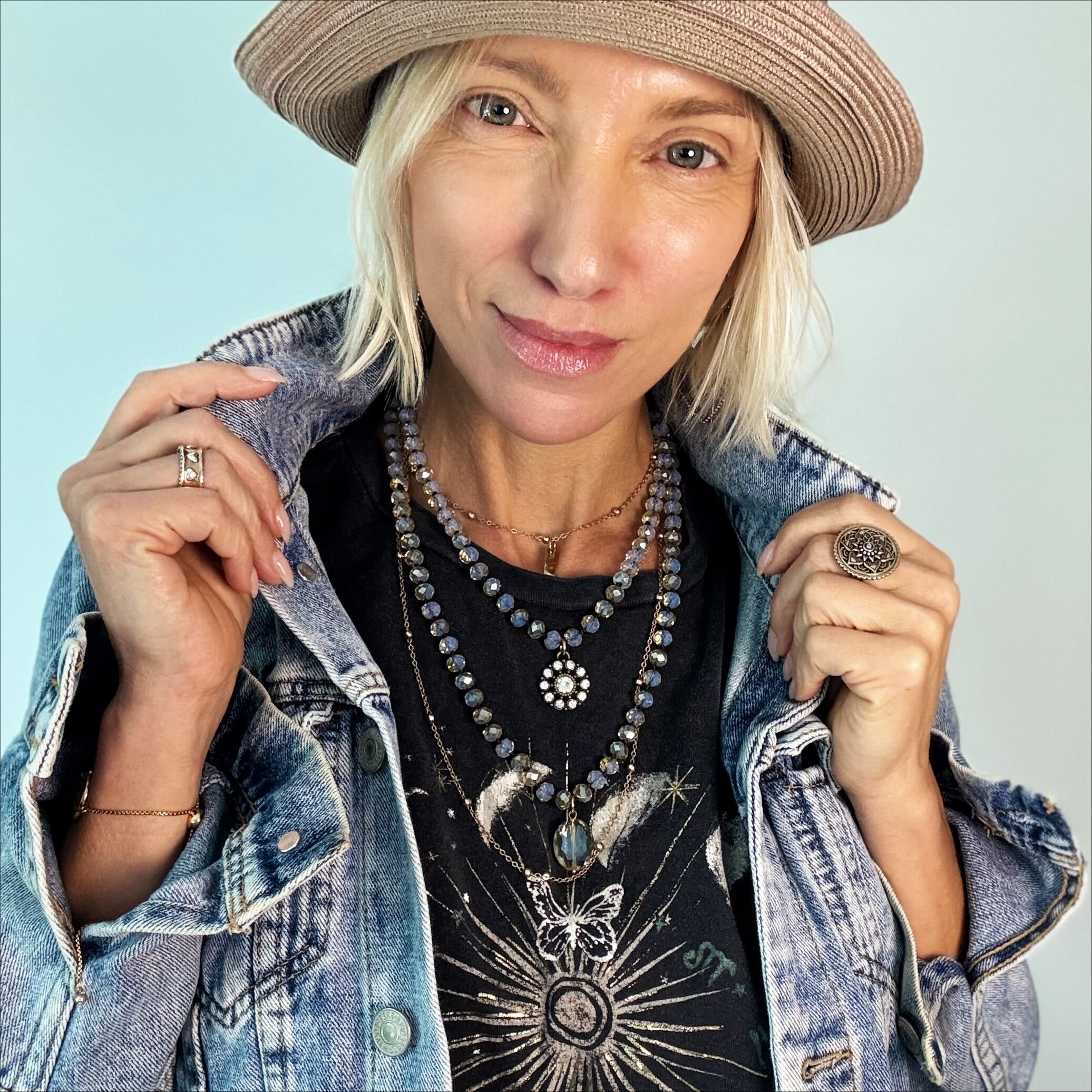 Woman wearing a denim jacket, hat, and layered necklaces against a light blue background