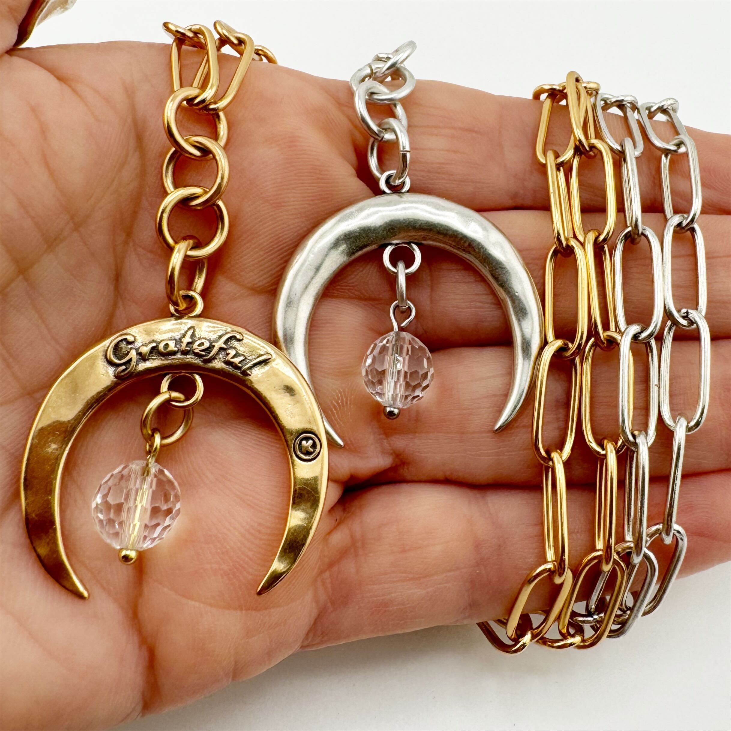 Two necklaces, one with a gold crescent moon pendant and the other with a silver crescent moon pendant, each accompanied by a faceted crystal quartz stone, displayed on a hand.