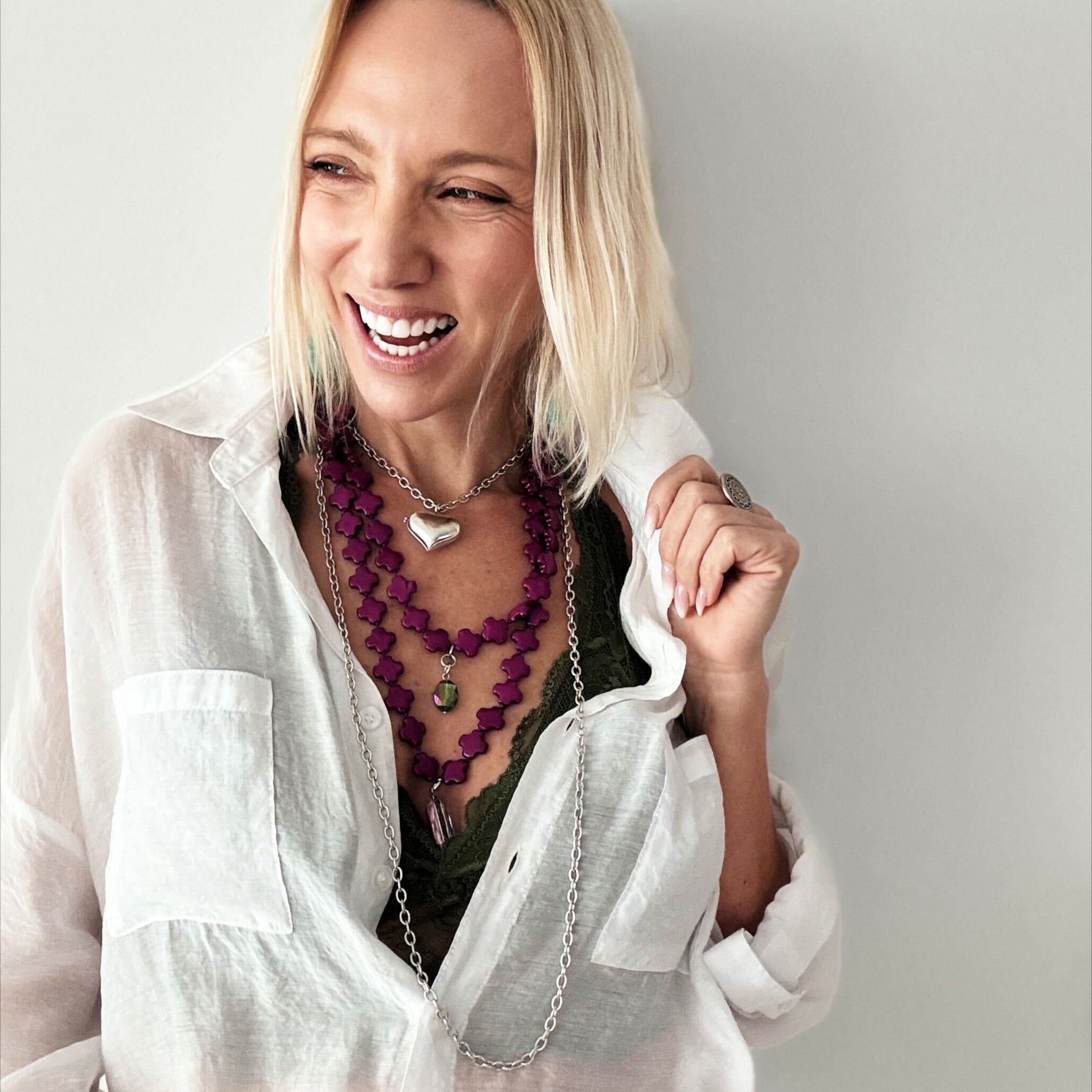 A person with blonde hair dons layered necklaces, showcasing a magenta purple howlite bead necklace and the heart locket essential in silver over a dark green top paired with a white shirt.