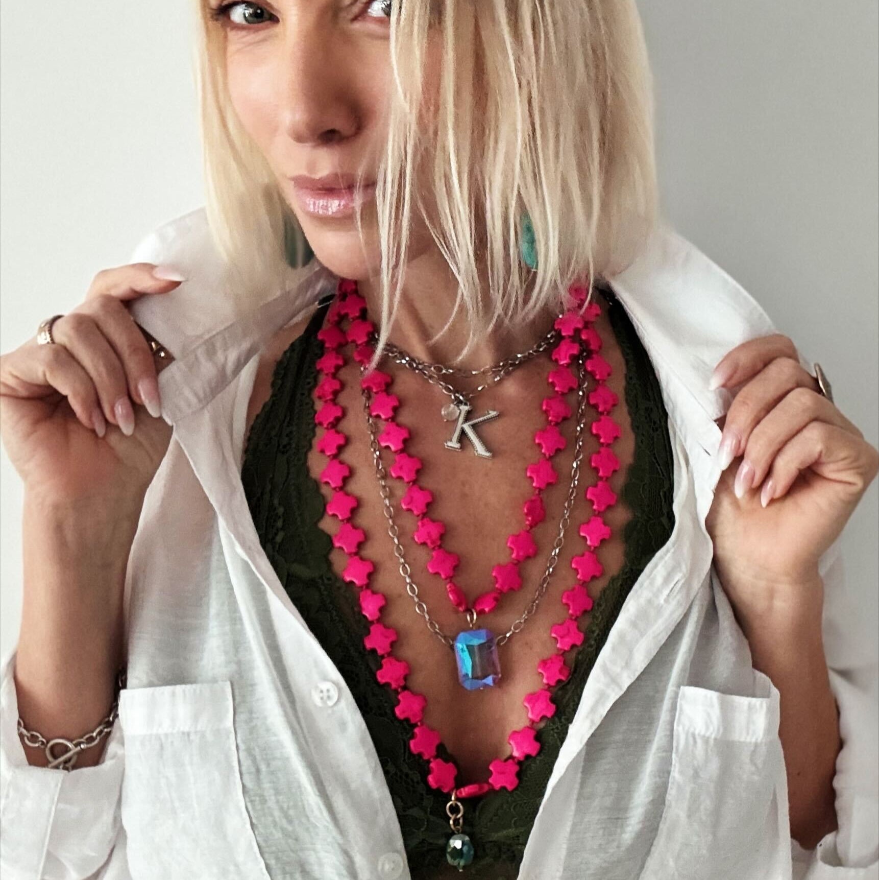 A person with blonde hair dons layered necklaces, showcasing a hot pink howlite bead necklace and a metallic "K" pendant, over a dark green top paired with a white shirt.