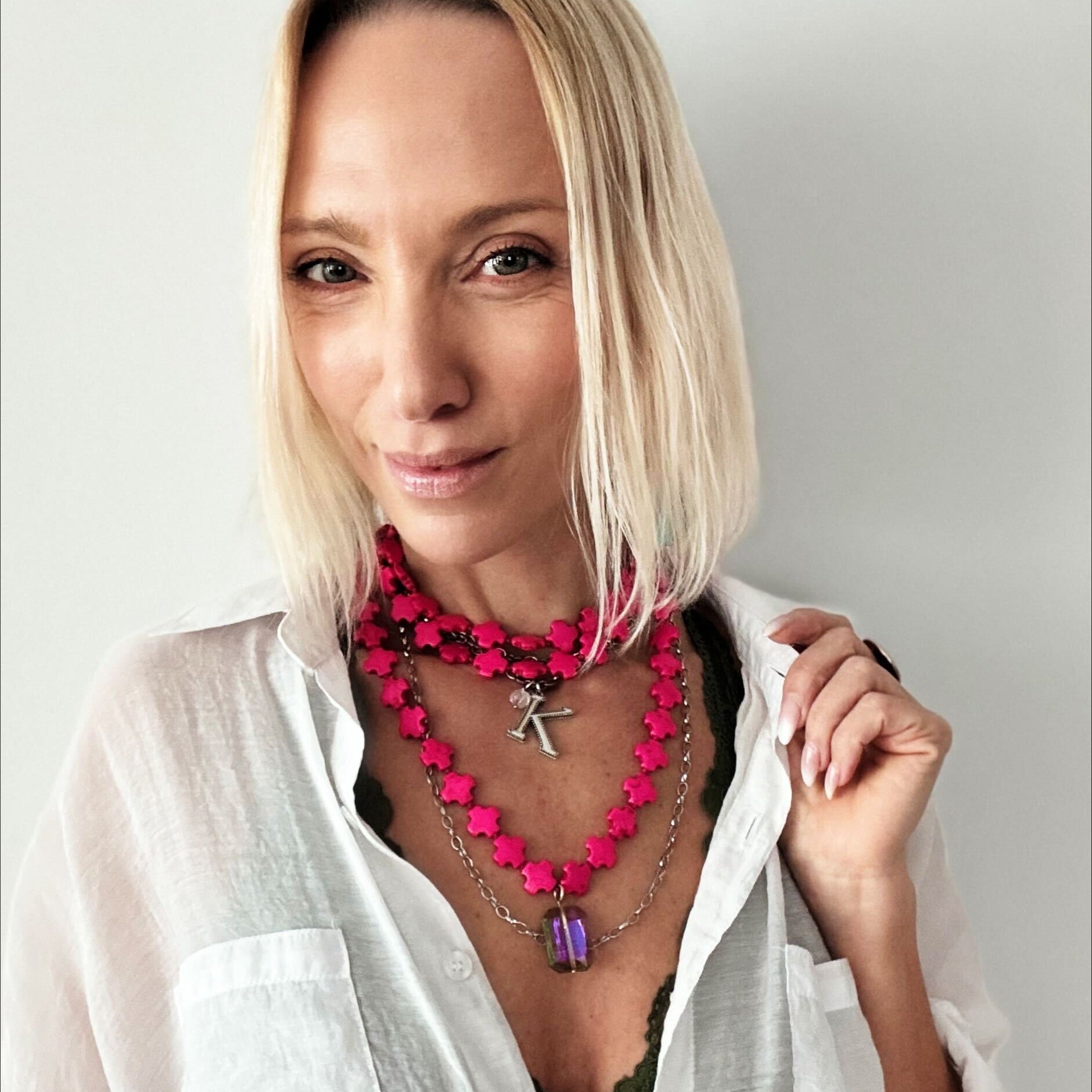 A person with blonde hair dons layered necklaces, showcasing a hot pink howlite bead necklace and a metallic "K" pendant, over a dark green top paired with a white shirt.