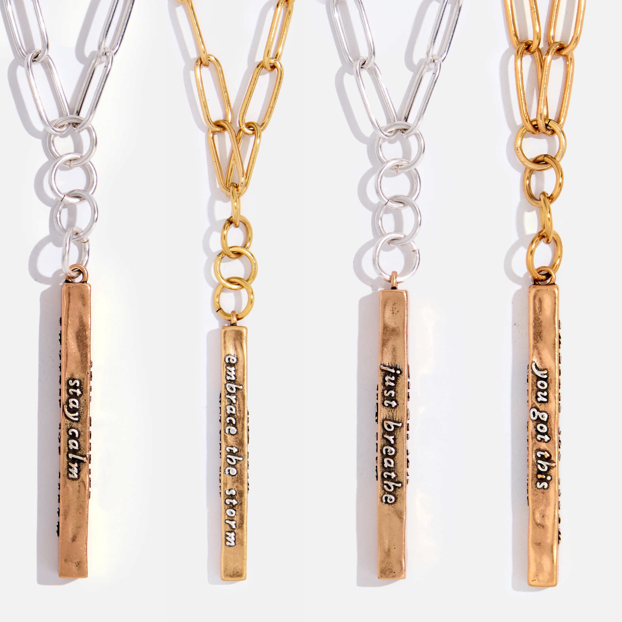 A collection of four 'Stay Calm' Klink necklaces, featuring plated brass paperclip chains and antique gold finish vertical bar pendants with messages on each side. Each necklace has a different length.