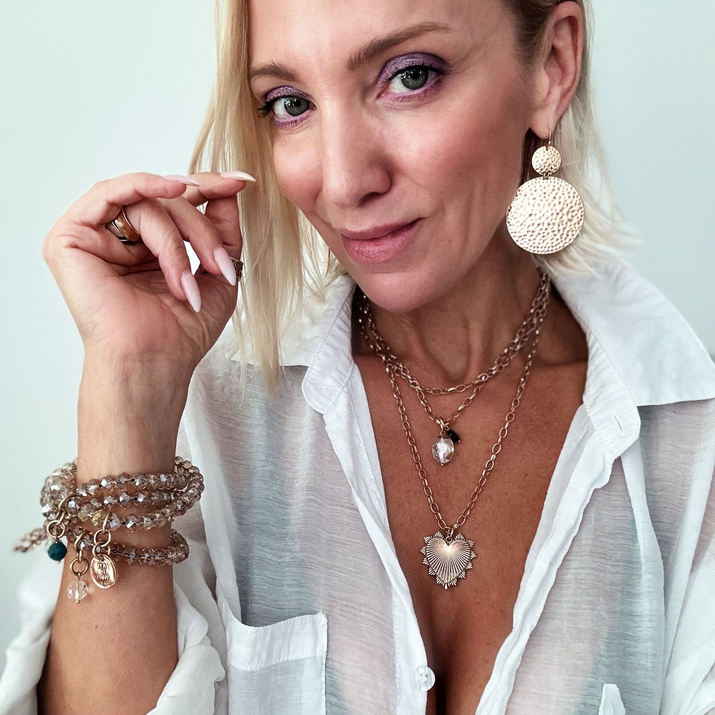 A person wearing a white shirt, showcasing multiple pieces of jewelry including a heart-shaped pendant necklace, a chain necklace with a faceted crystal accent, and several bracelets on the wrist.