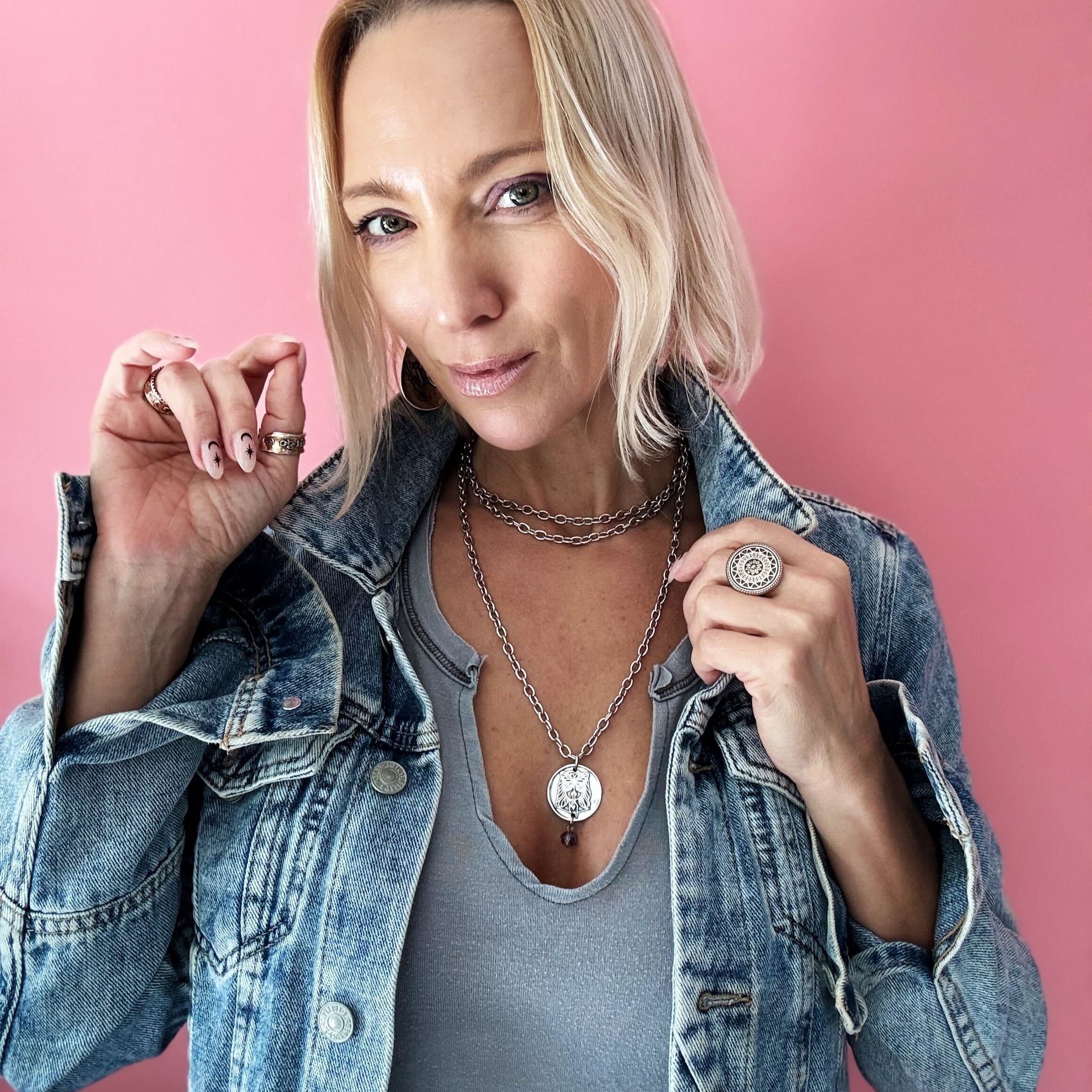 Person in a denim jacket and gray shirt poses against a pink background, showcasing layered necklaces with a chain necklace and rings shimmering with crystal accents.
