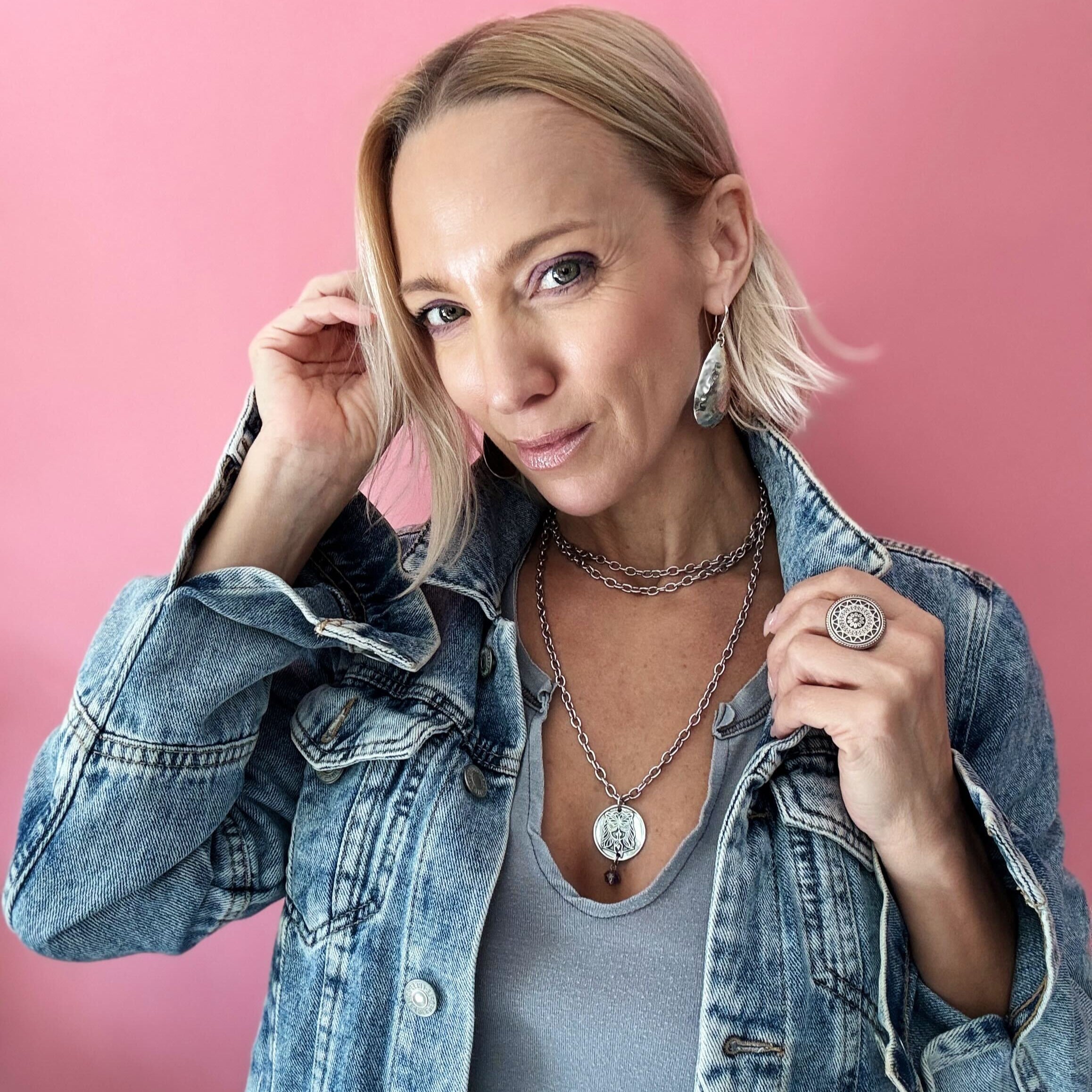 Person in a denim jacket and gray shirt poses against a pink background, showcasing layered necklaces with a chain necklace and rings shimmering with crystal accents.