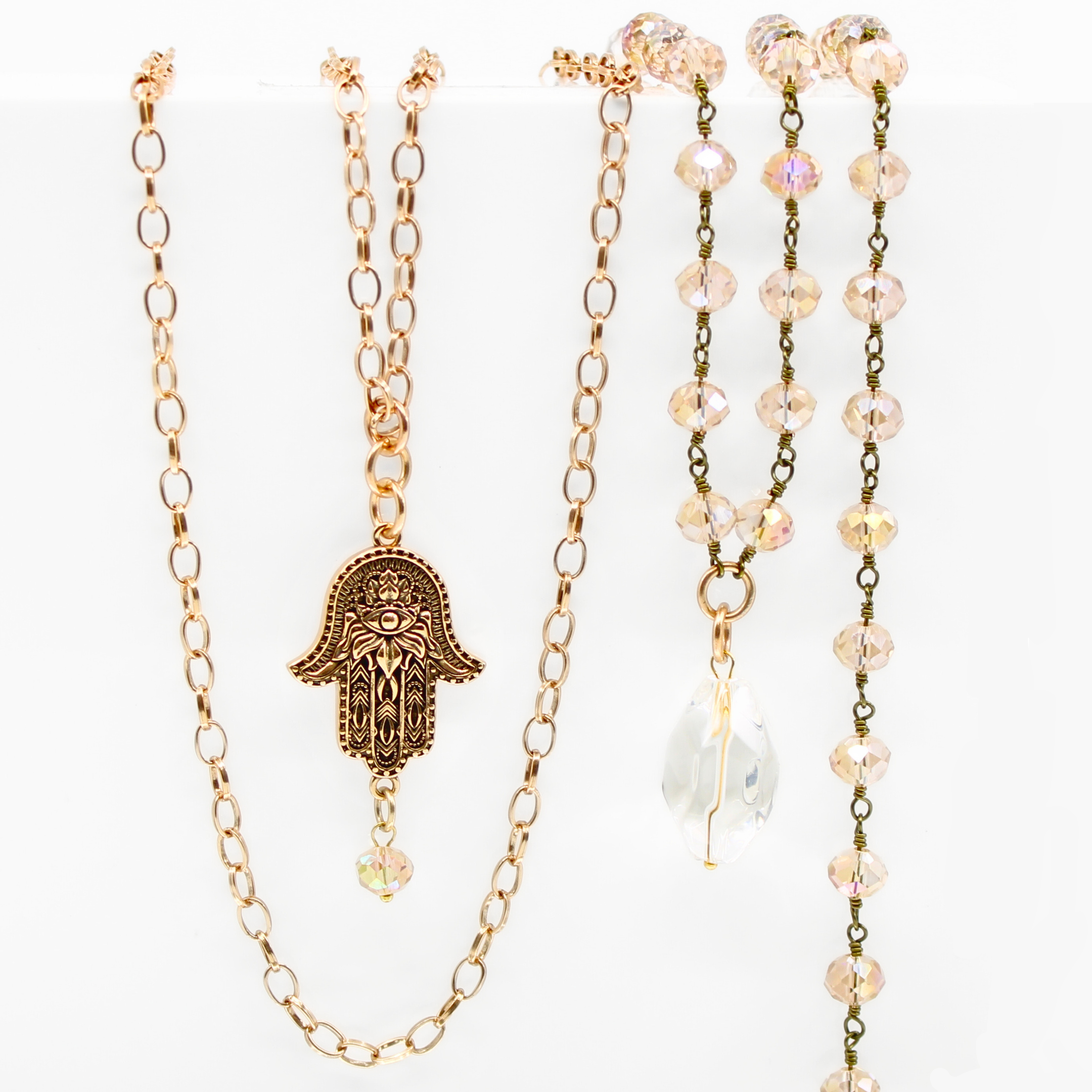A collection of layered rosary necklaces with beige crystal beads, metallic chains, and decorative pendants including a peace symbol and a hand of Hamsa.