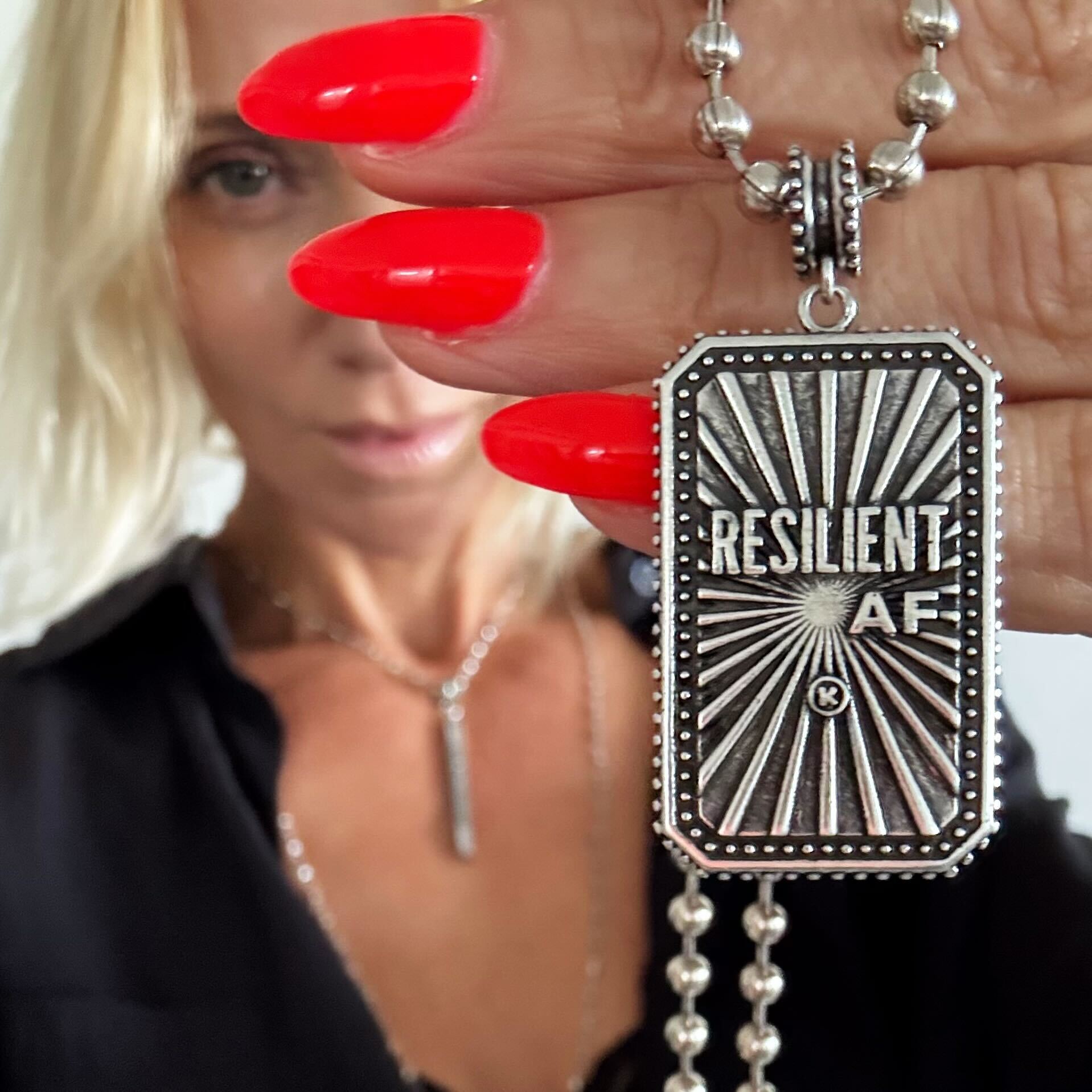 Silver pendant with 'RESILIENT AF' text held by a person with red nails.