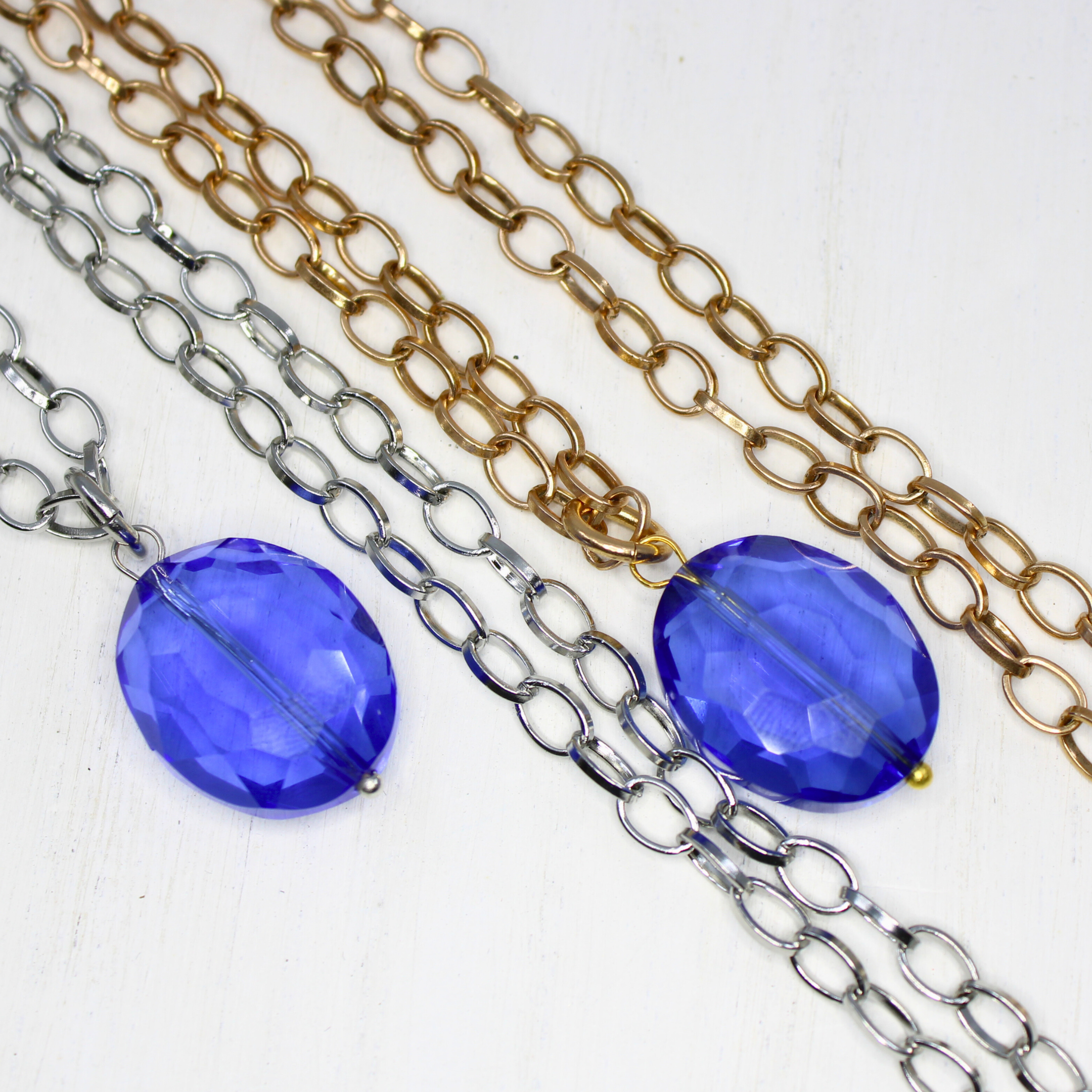 Two chain necklaces, one with a silver finish and the other with an antique gold finish, each featuring a large faceted royal blue crystal.