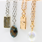 Three necklaces with varying pendants. From left to right: a silver chain with a rectangular pendant that reads 'Breathe', a gold chain with a heart-shaped pendant, and a third with a clear crystal pendant.