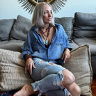 Woman sitting on a couch wearing a blue denim shirt and ripped jeans, with a decorative wall mirror in the background.