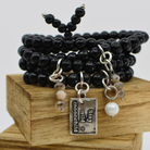 A person's wrist wearing multiple black wooden bead wrap bracelets with gold and silver colored pendants, and faceted crystal accents.