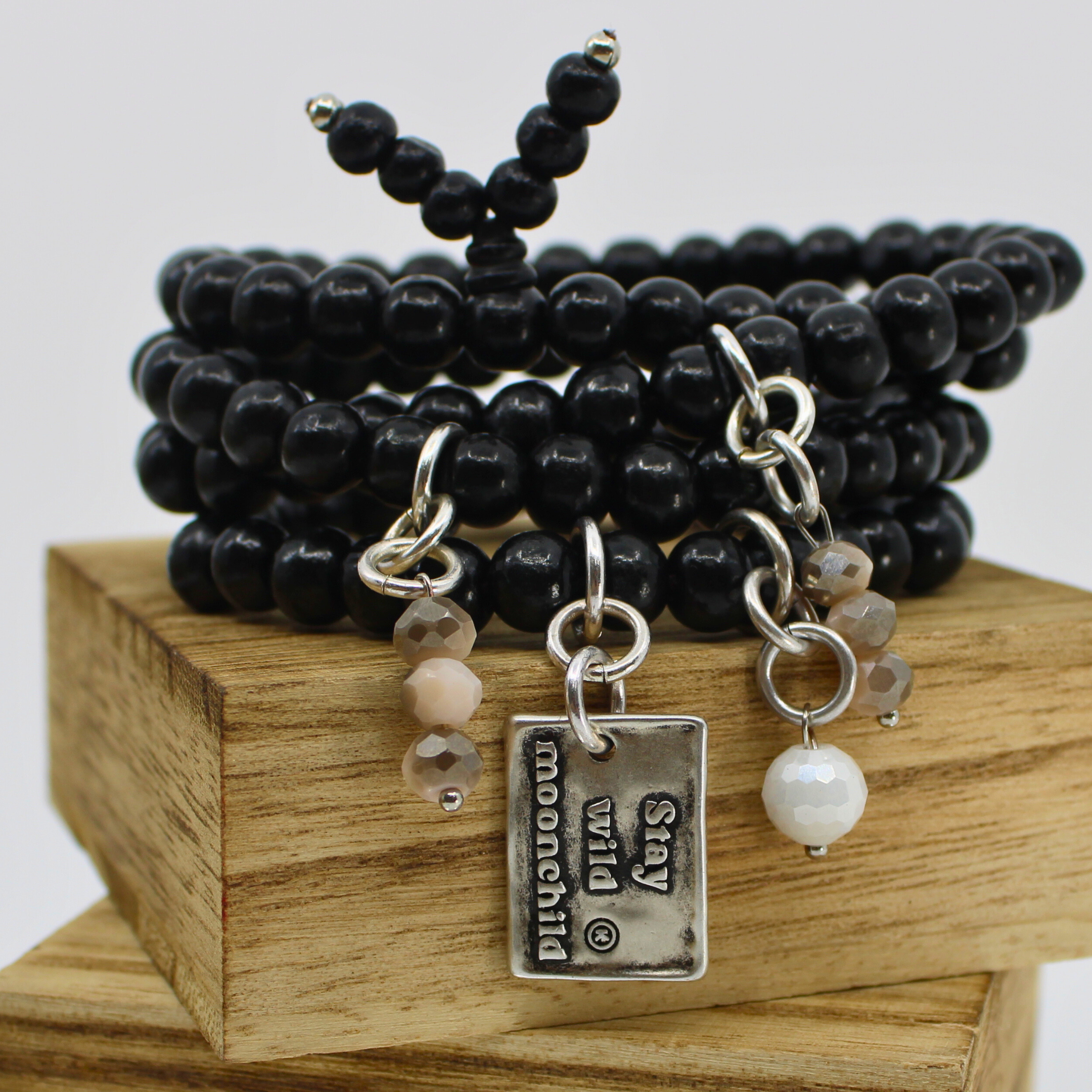 A person's wrist wearing multiple black wooden bead wrap bracelets with gold and silver colored pendants, and faceted crystal accents.