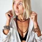 Woman wearing layered necklaces and bracelets with a white shirt on a plain background