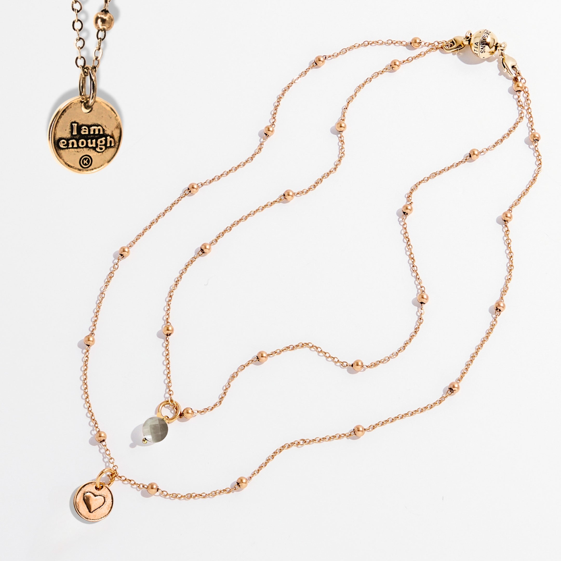 Two dainty necklaces with pendants, one with a silver finish and the other with a gold finish. Both have a chain link design and feature a circular pendant with the phrase 'I am enough' and a heart symbol respectively.