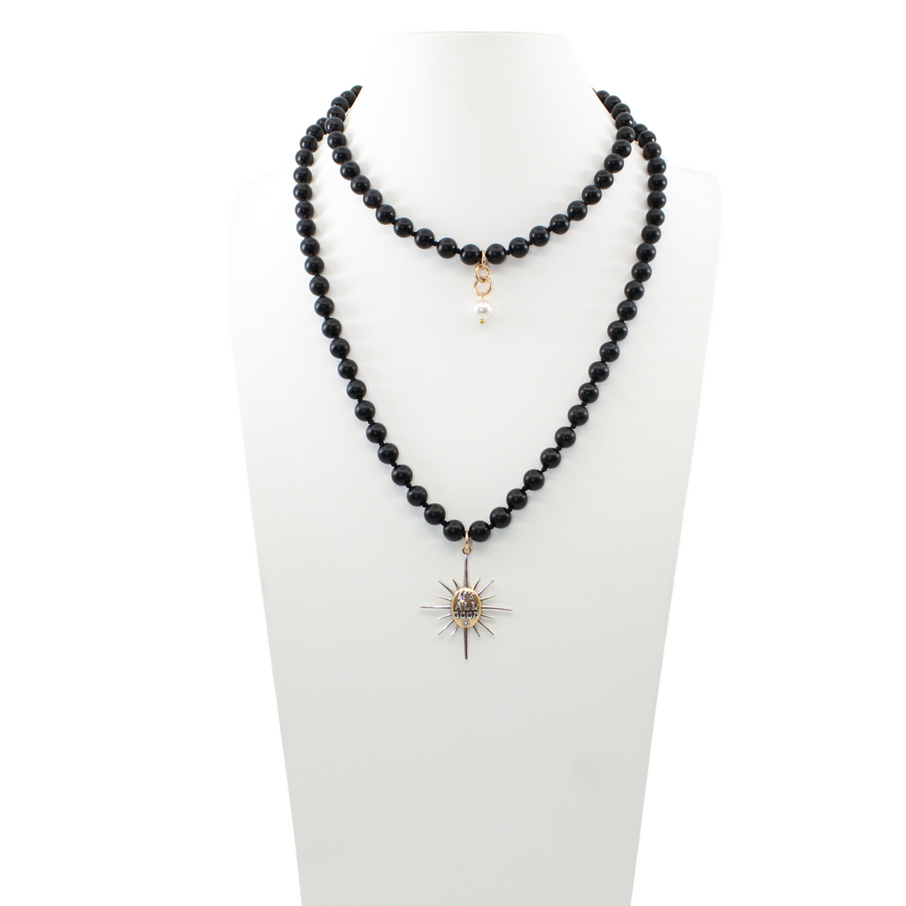 Black onyx necklace with a sunburst pendant reading Its All Good. Gold clasp and pearl accent detail on chain.