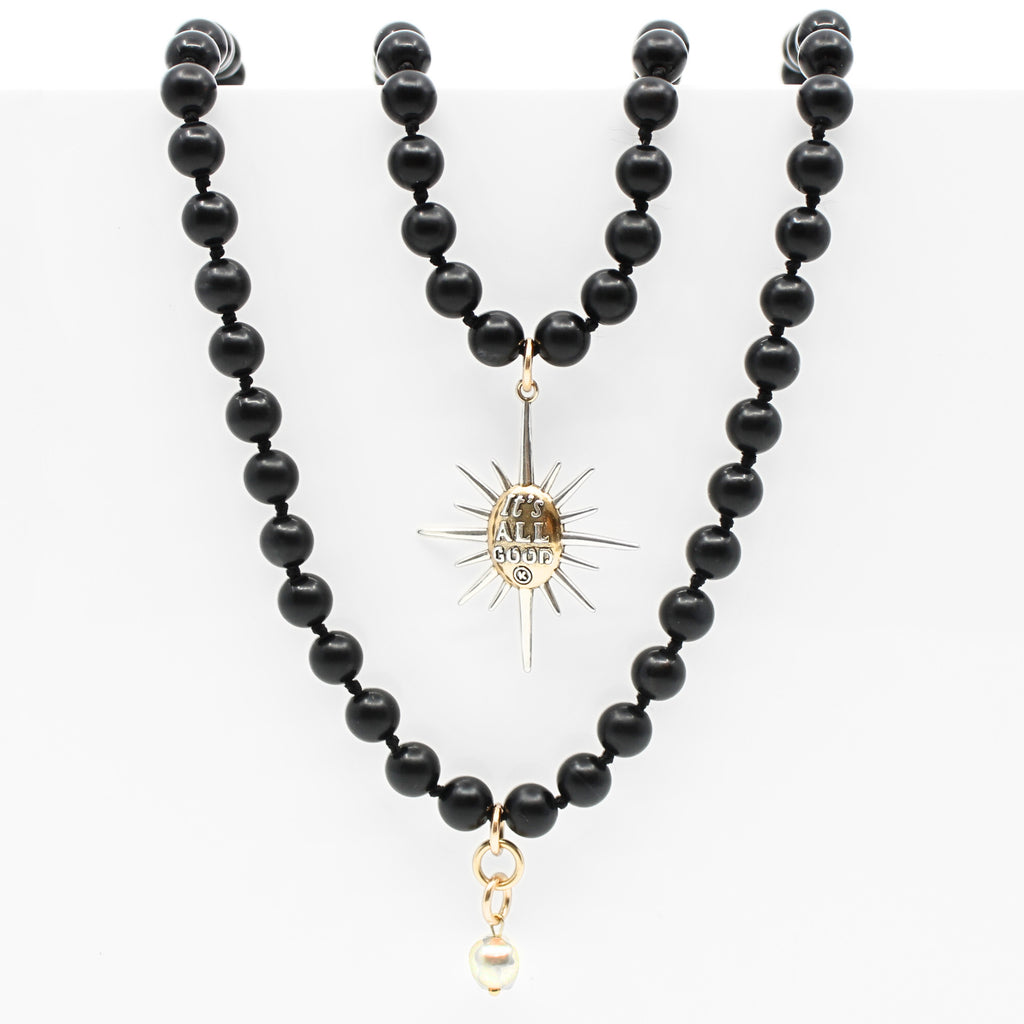 Black onyx necklace with a sunburst pendant reading Its All Good. Gold clasp and pearl accent detail on chain.