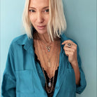 Woman in teal shirt with tousled blonde bob, wearing a butterfly pendant and large ring, against a blue background.