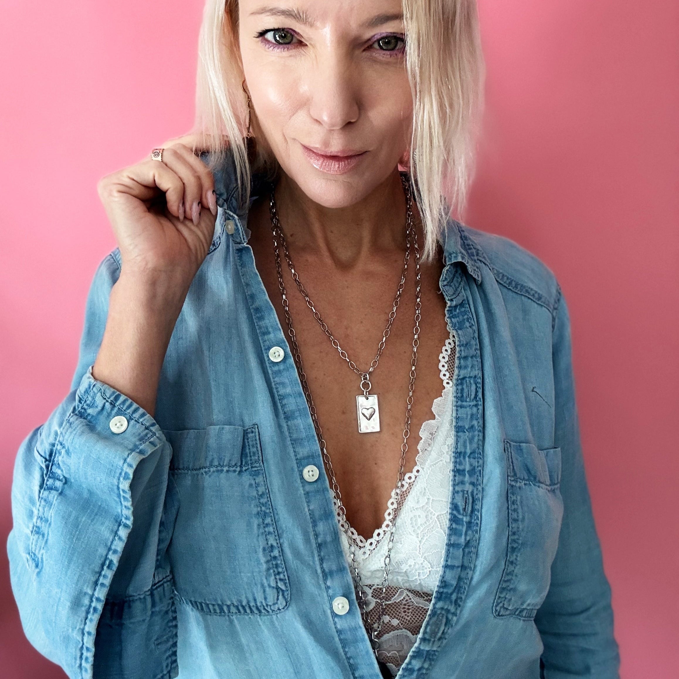 A person wearing a denim shirt and a white lace top, showcasing a silver-colored stainless steel chain necklace with a square pendant.