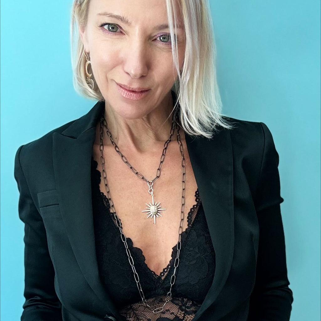 A woman in a black blazer smiles, adorned with layered necklaces featuring a gunmetal paperclip chain and sunburst pendant, along with a large ring, against a blue background.