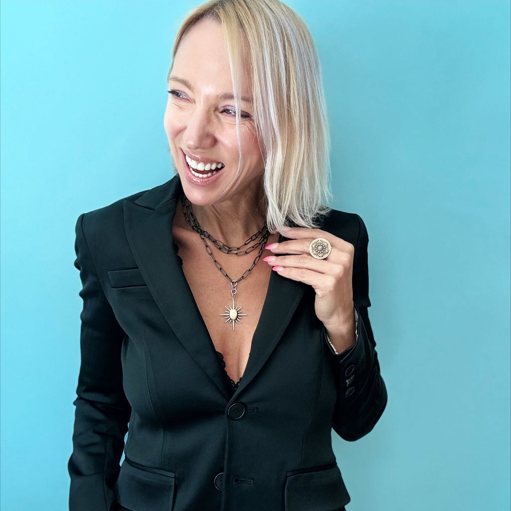 A woman in a black blazer smiles, adorned with layered necklaces featuring a gunmetal paperclip chain and sunburst pendant, along with a large ring, against a blue background.